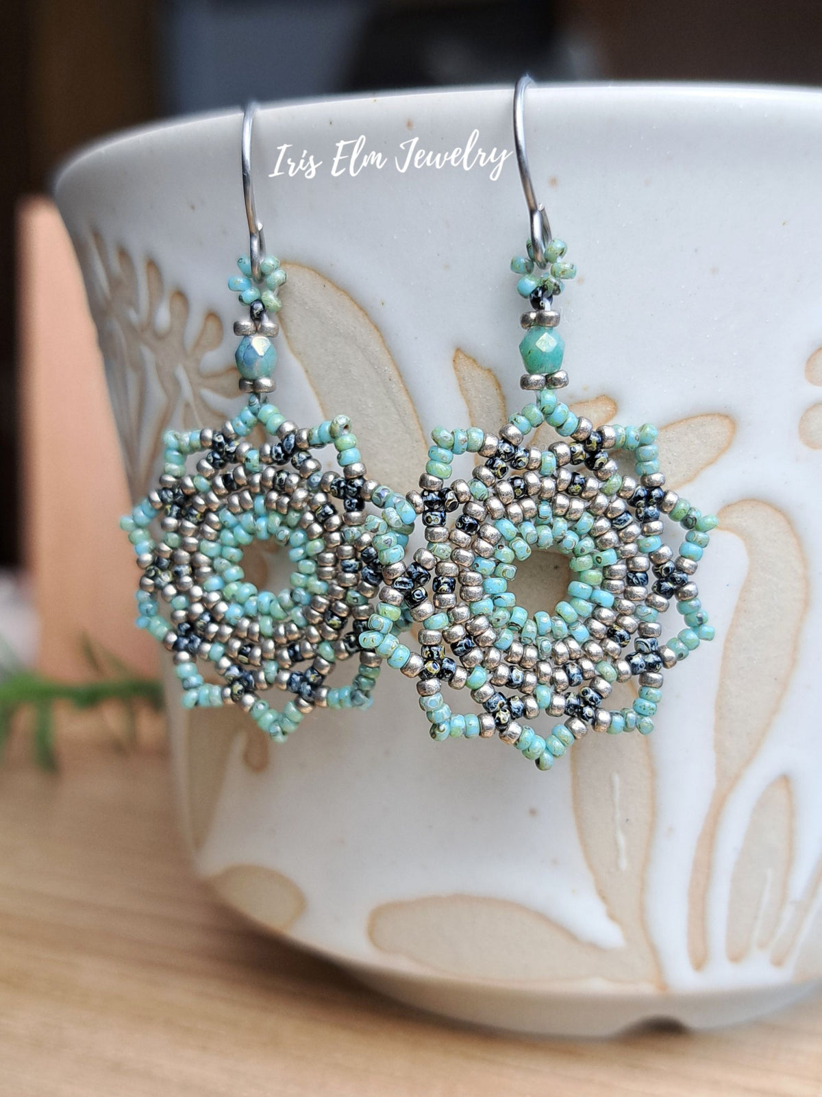 A pair of handmade beaded dangle earrings with intricate geometric patterns in light blue, silver, and black seed beads, hanging from silver fishhook wires. The earrings are displayed in front of a white ceramic mug with beige floral patterns on a wooden surface.