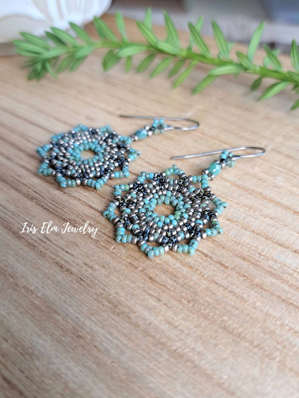 A pair of handmade beaded dangle earrings with intricate spiral patterns in turquoise, black, and silver, resting on a light wood surface with a green sprig in the background. The brand name 'Iris Elm Jewelry' is watermarked on the left.