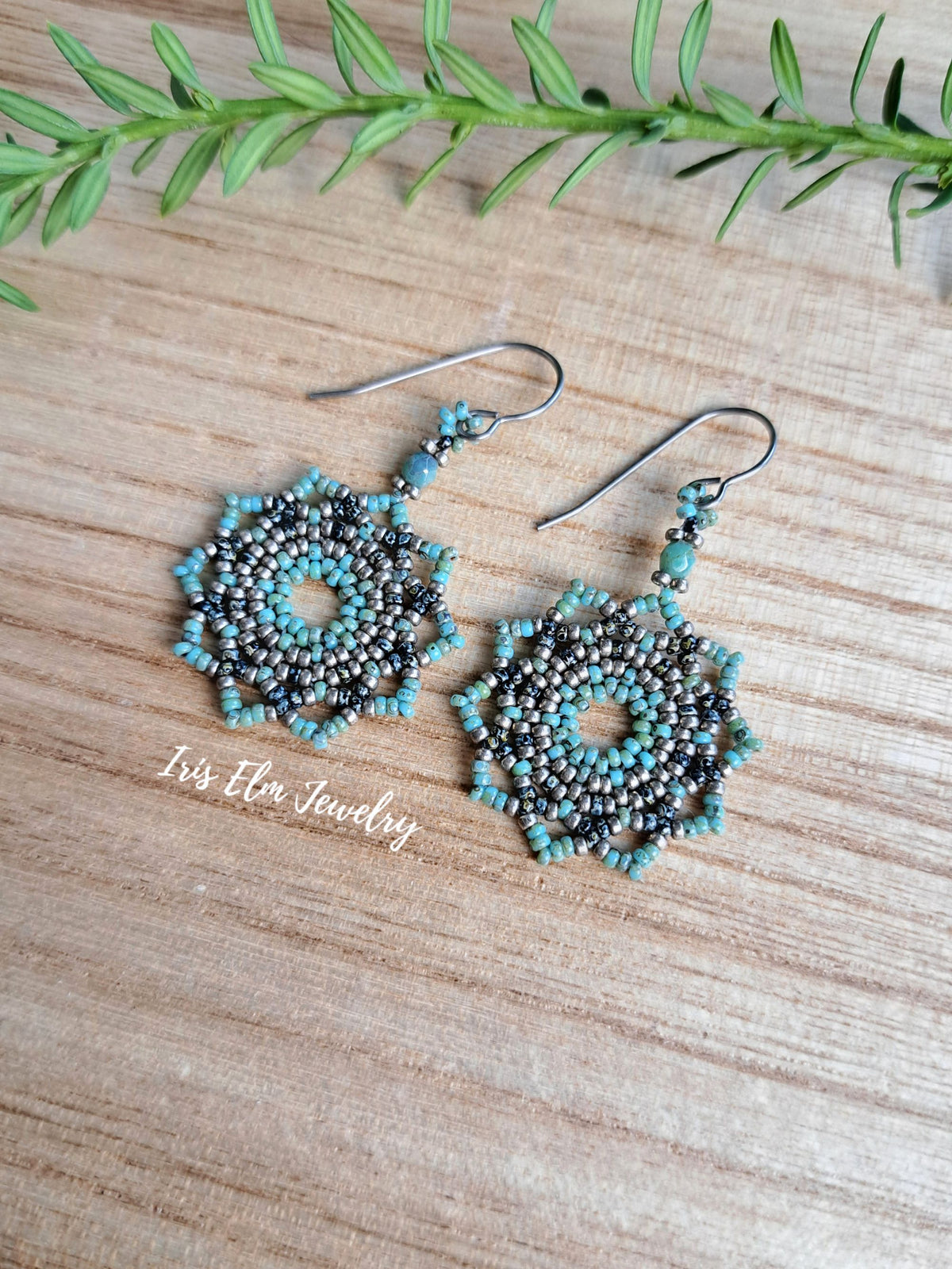 A pair of handmade beaded dangle earrings with turquoise and dark gray beads, displayed on a light wood surface with a green sprig in the background. The brand name 'Iris Elm Jewelry' is printed on the image.