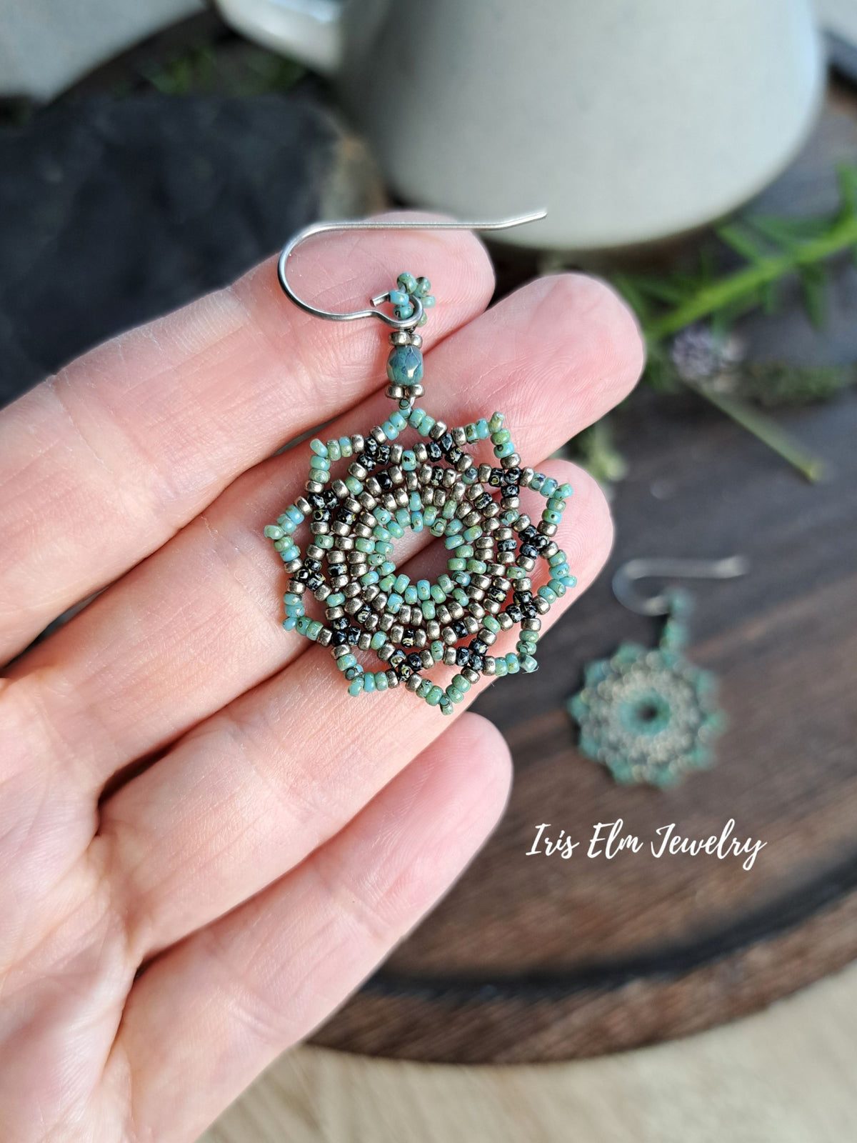 A hand holds a pair of intricate beaded dangle earrings with silver fishhook wires. The earrings feature a circular mandala-like design made of turquoise, dark bronze, and silver seed beads, with a small turquoise bead at the top.