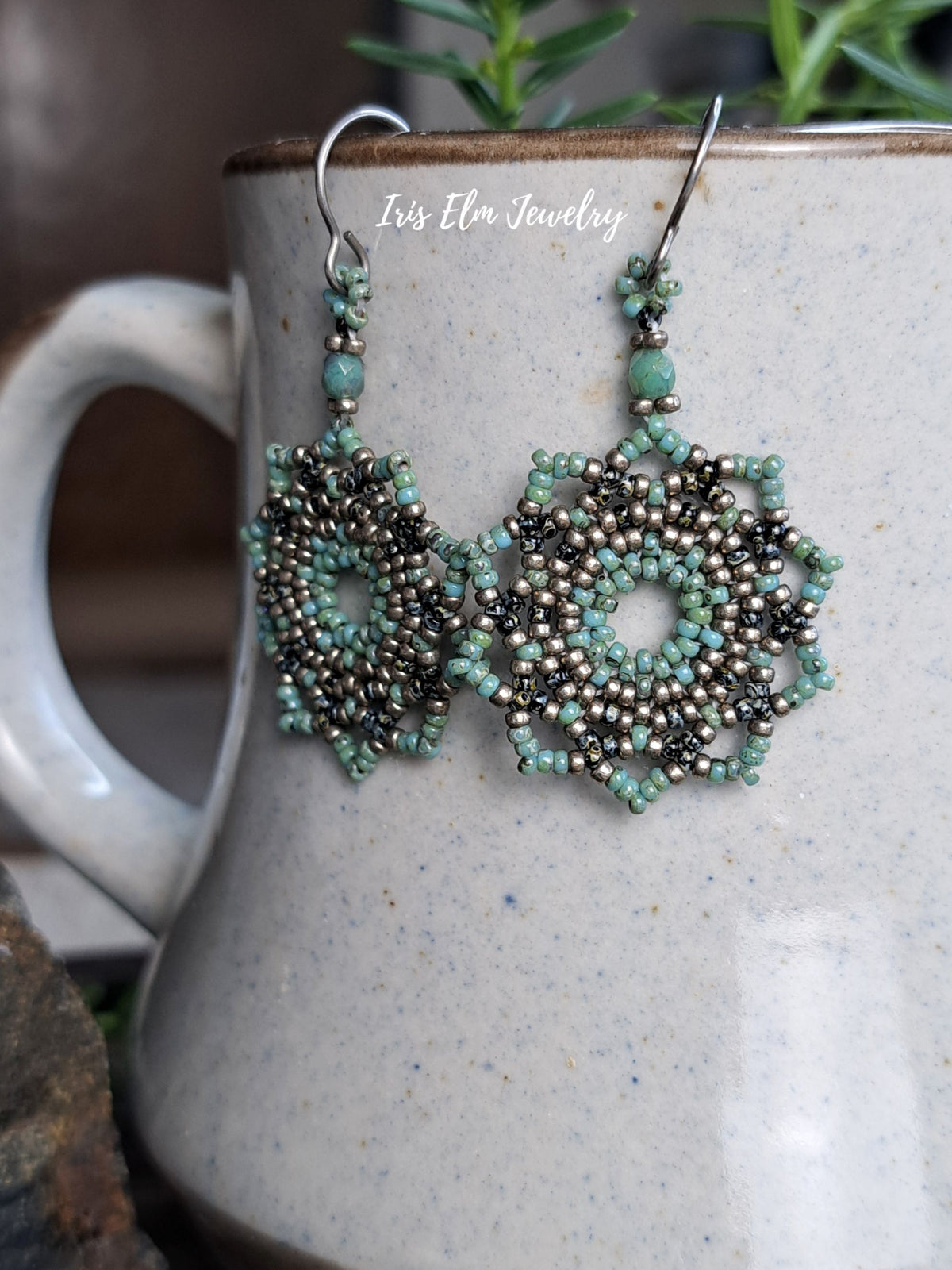 A pair of handmade beaded dangle earrings with intricate floral patterns in turquoise, black, and metallic silver, hanging from silver fishhook wires. The earrings are displayed against a speckled beige ceramic mug with a green plant in the background.