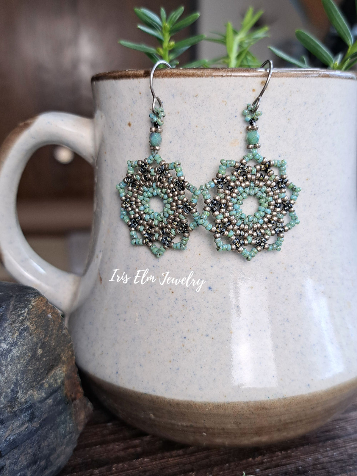 A pair of handmade beaded dangle earrings with intricate floral/starburst patterns in turquoise, dark brown, and silver, hanging from silver fishhook wires. The earrings are displayed draped over a speckled beige ceramic mug with green sprigs in the background.
