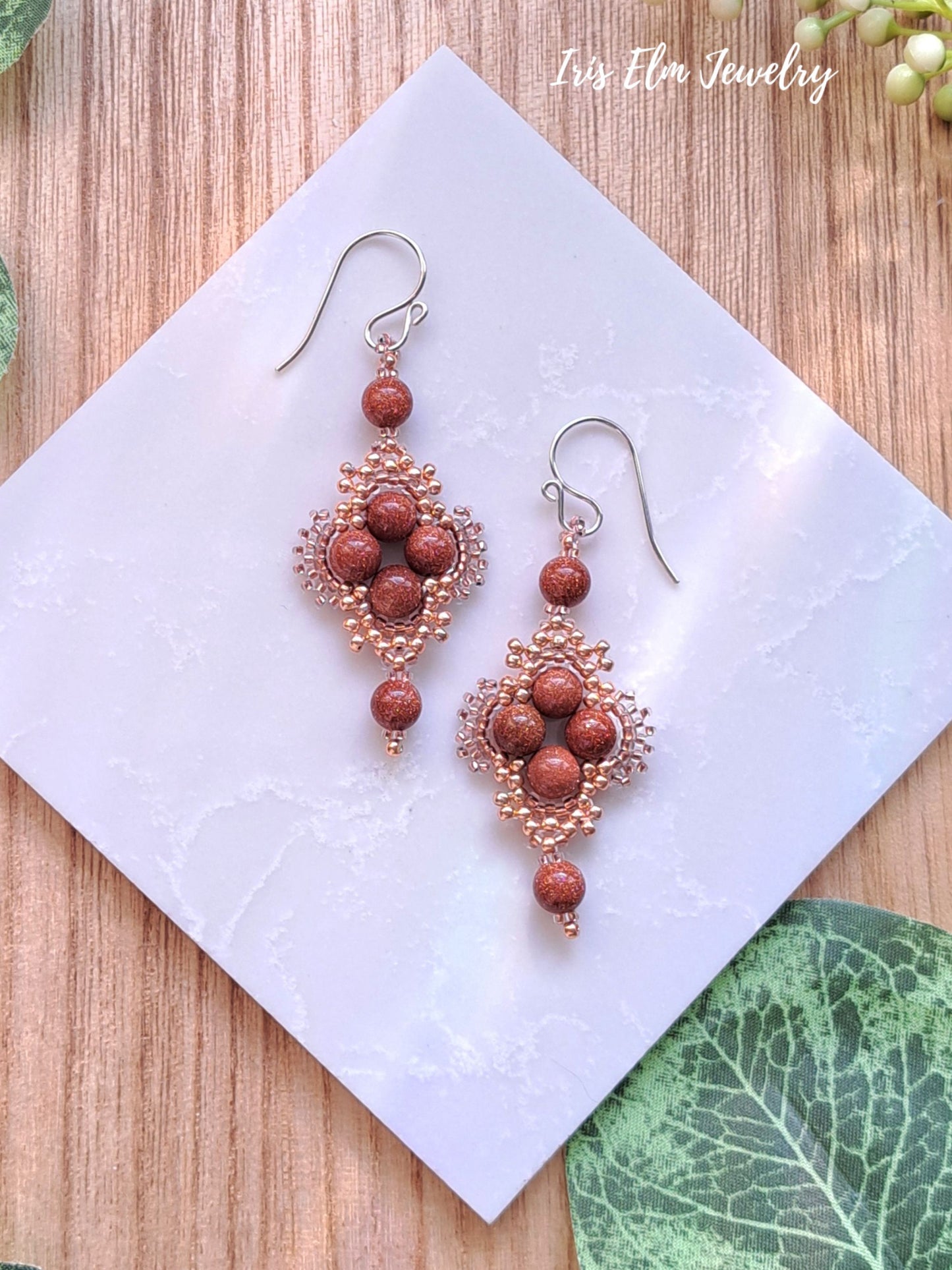 Pair of beaded brown goldstone beadwoven earrings on a white square with 'Iris Elm Jewelry' branding.