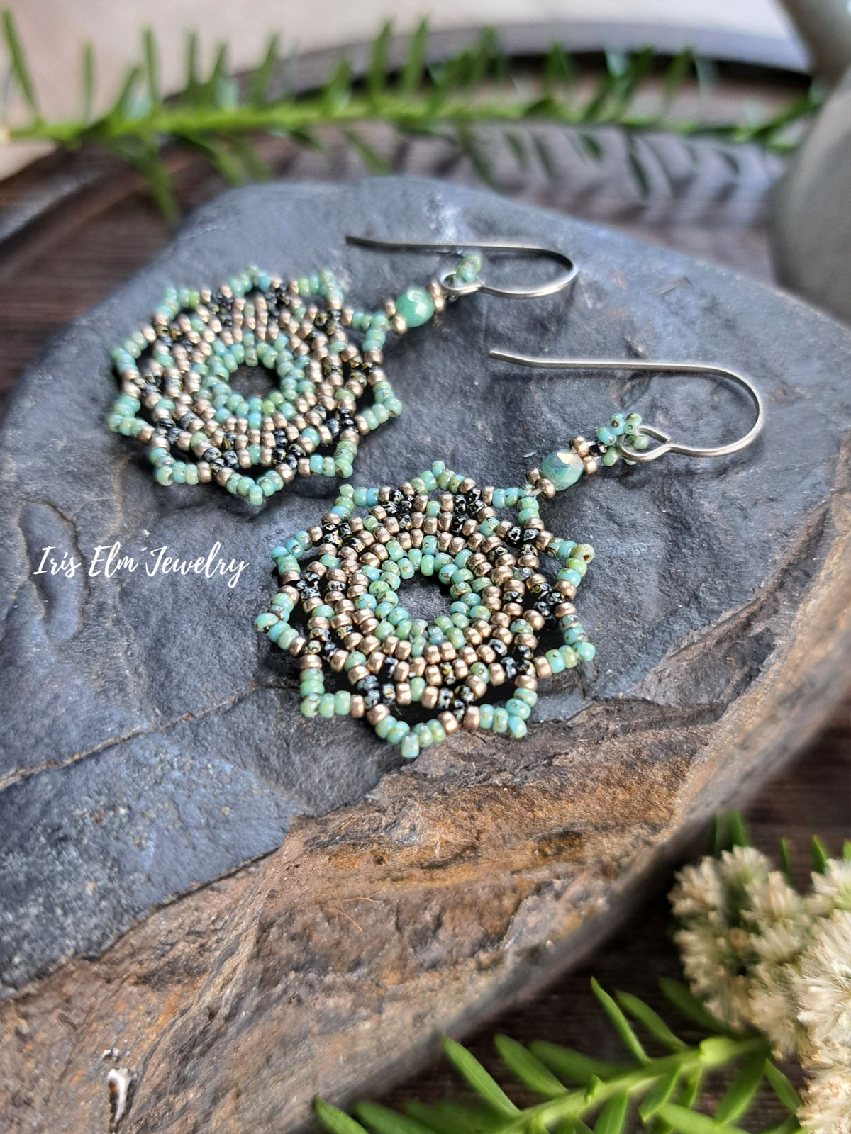 A pair of handmade beaded dangle earrings with silver fishhook wires, featuring intricate spiral patterns of mint green, black, and metallic gold seed beads. The earrings are displayed on a textured dark gray stone with green foliage and small white flowers in the background, with a watermark reading 'Iris Elm Jewelry' on the left.