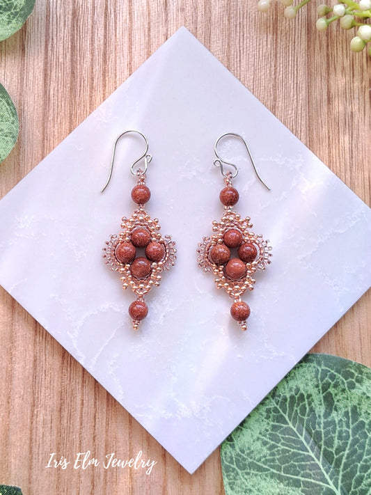 Ornate earrings with brown goldstone and copper color seed beads on a white square pad, brand 'Iris Elm Jewelry' visible.