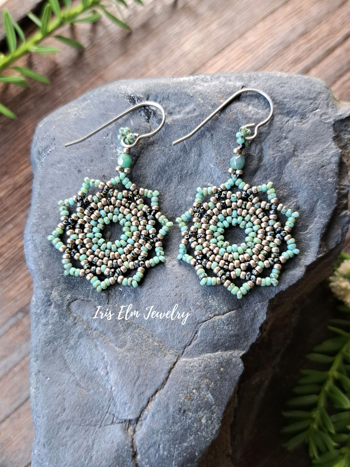 A pair of handmade beaded dangle earrings with a geometric starburst design, displayed on a dark gray stone. The earrings feature light blue-green beads, black seed beads, and metallic bronze beads on silver-toned French hook ear wires.