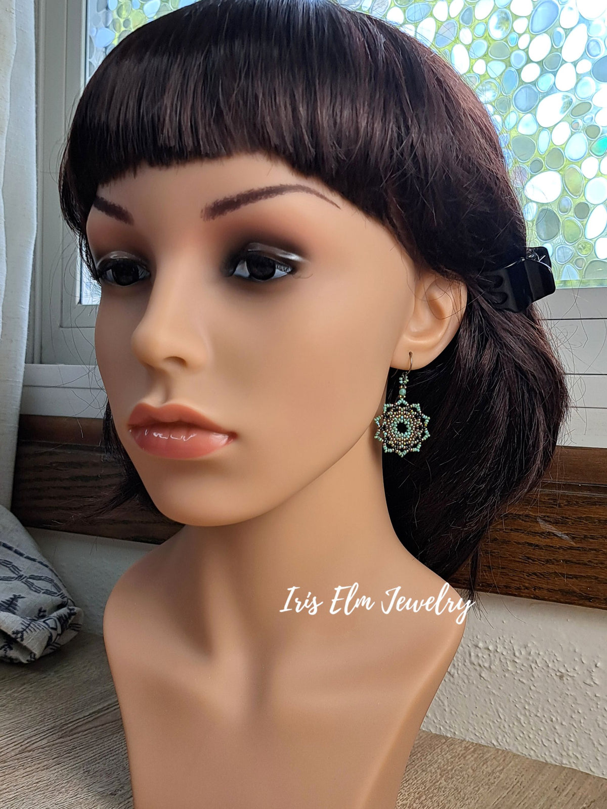 A mannequin bust wearing a dangling earring with a green and silver floral/starburst design. The mannequin has dark brown hair and is photographed indoors near a window, with the text 'Iris Elm Jewelry' overlaid near the lower right.