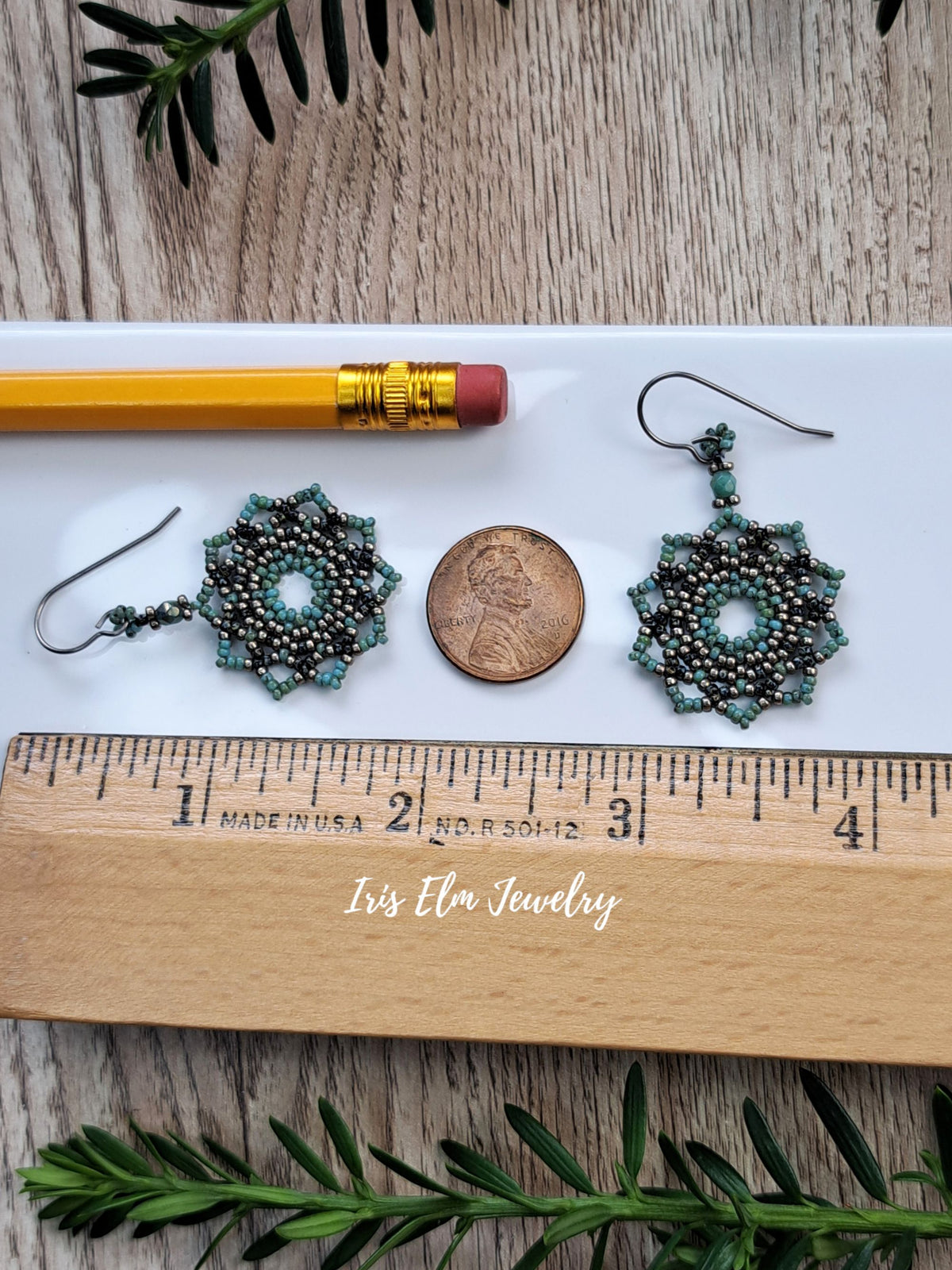 A pair of 1.75 inch long handmade beaded earrings with a starburst design in turquoise, green, and black beads, displayed on a white tray with a pencil, a penny, and a wooden ruler for scale. The tray has 'Iris Elm Jewelry' written on it, and the background is a rustic wood surface with green sprigs.