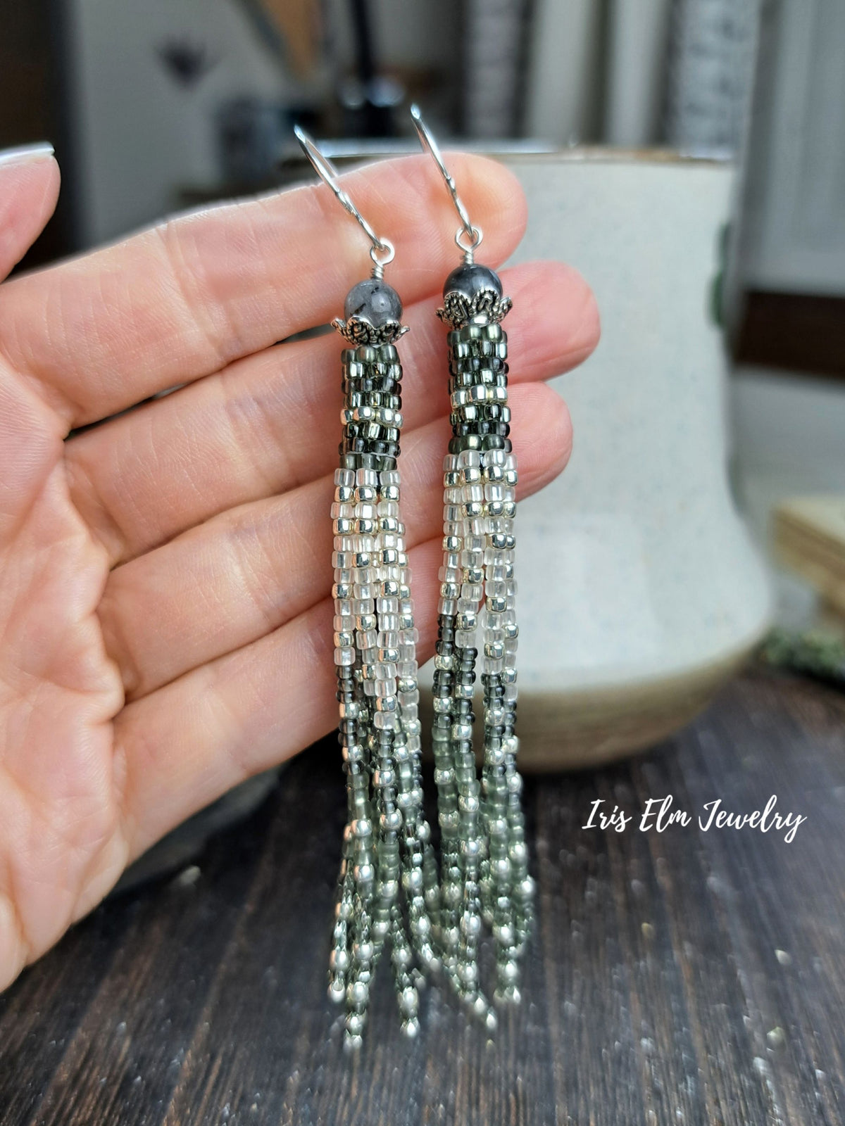 A hand holds a pair of long beaded tassel earrings with silver fishhook wires. The earrings feature multiple strands of small beads in silver, gray, and dark metallic tones.