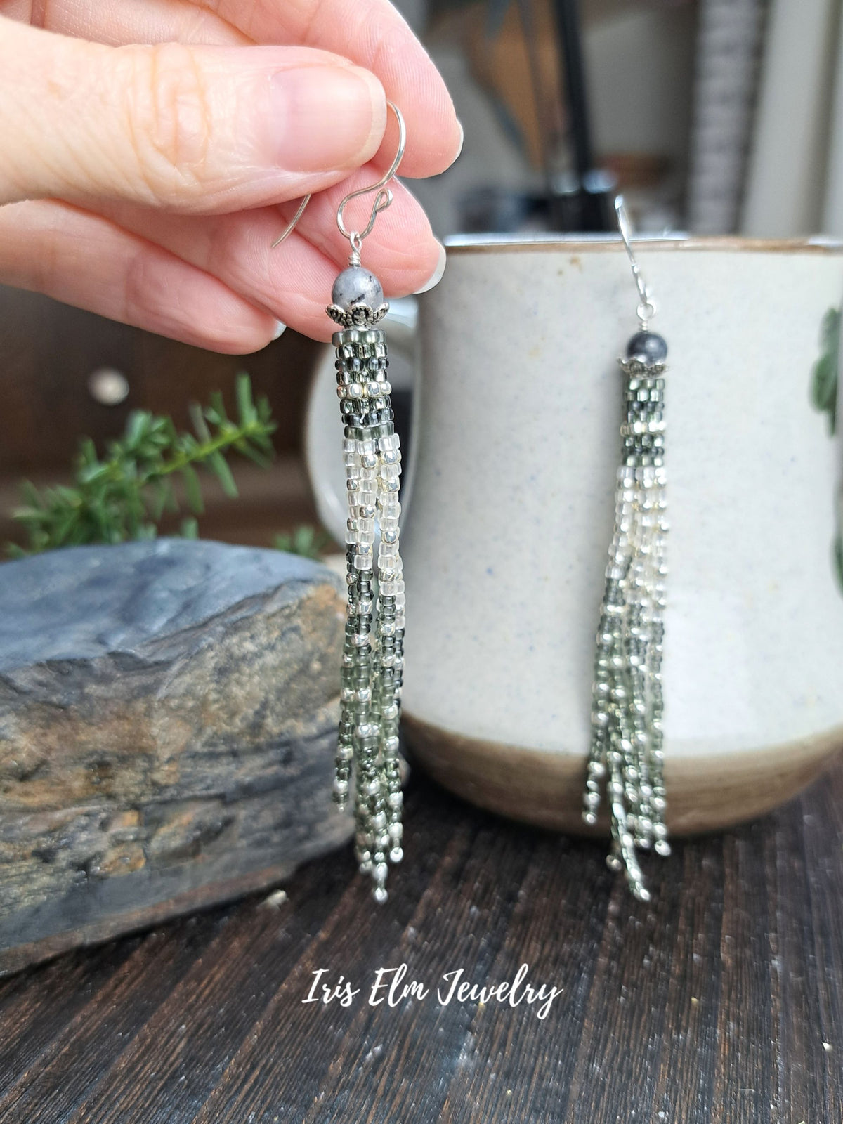 A hand holds a pair of dangling beaded earrings with silver-toned hooks and multiple strands of clear and green beads, with a ceramic mug and wooden surface in the background. The text 'Iris Elm Jewelry' appears at the bottom.