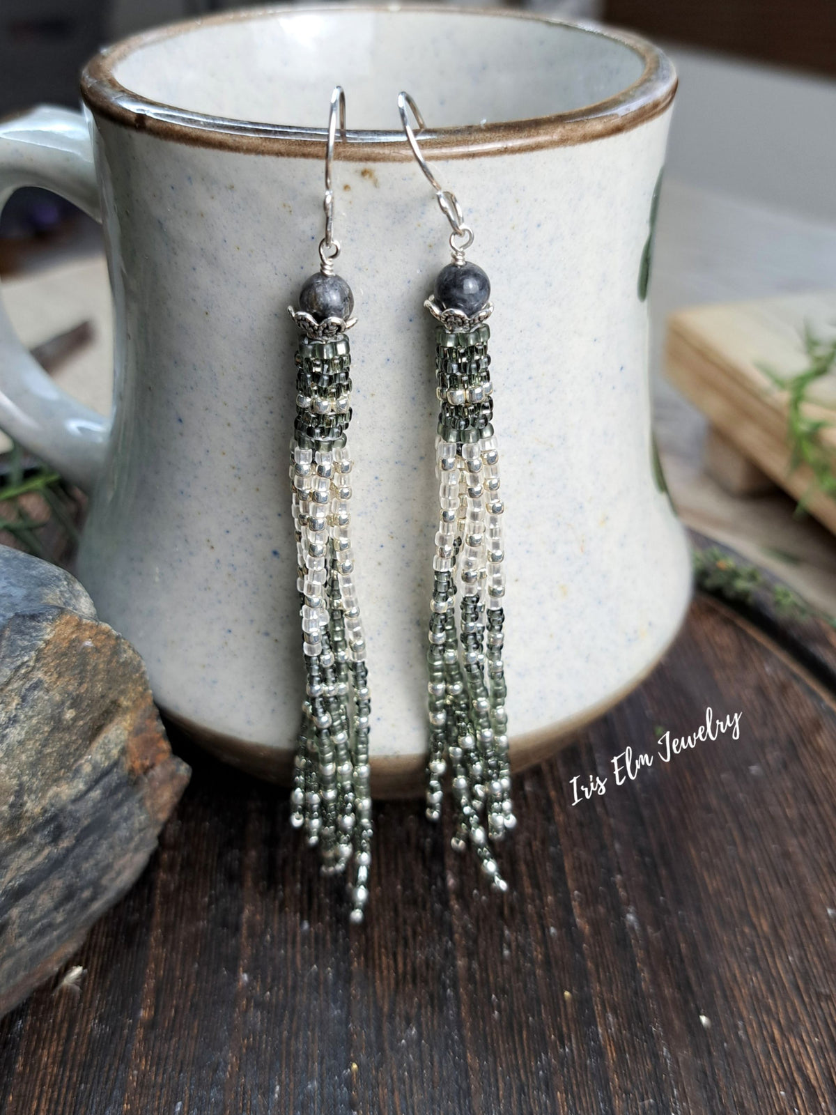 A pair of long beaded dangle earrings with dark gray beads and silver-toned chains, hanging from a ceramic mug on a rustic wooden surface. The text 'Iris Elm Jewelry' is visible on the right side.