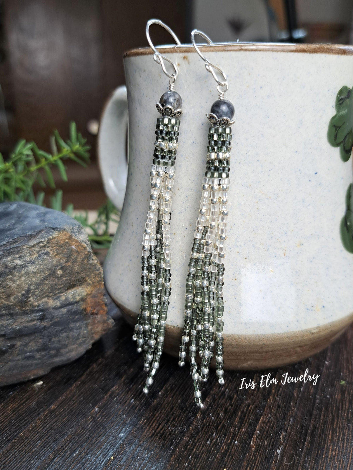 A pair of long beaded tassel earrings with silver fishhook wires and dark gray beads, hanging in front of a ceramic mug on a dark wooden surface. The earrings feature green and silver beads, with a watermark reading 'Iris Elm Jewelry' in the lower right corner.