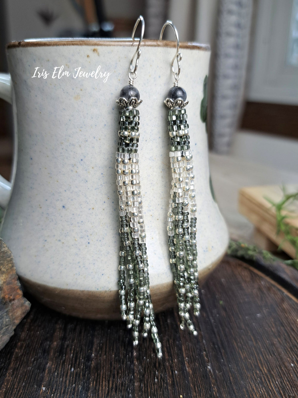 A pair of long beaded tassel earrings with silver-toned hooks, dark gray beads, and cascading green, silver, and clear beads, hanging in front of a speckled ceramic mug. The text 'Iris Elm Jewelry' is visible on the mug.