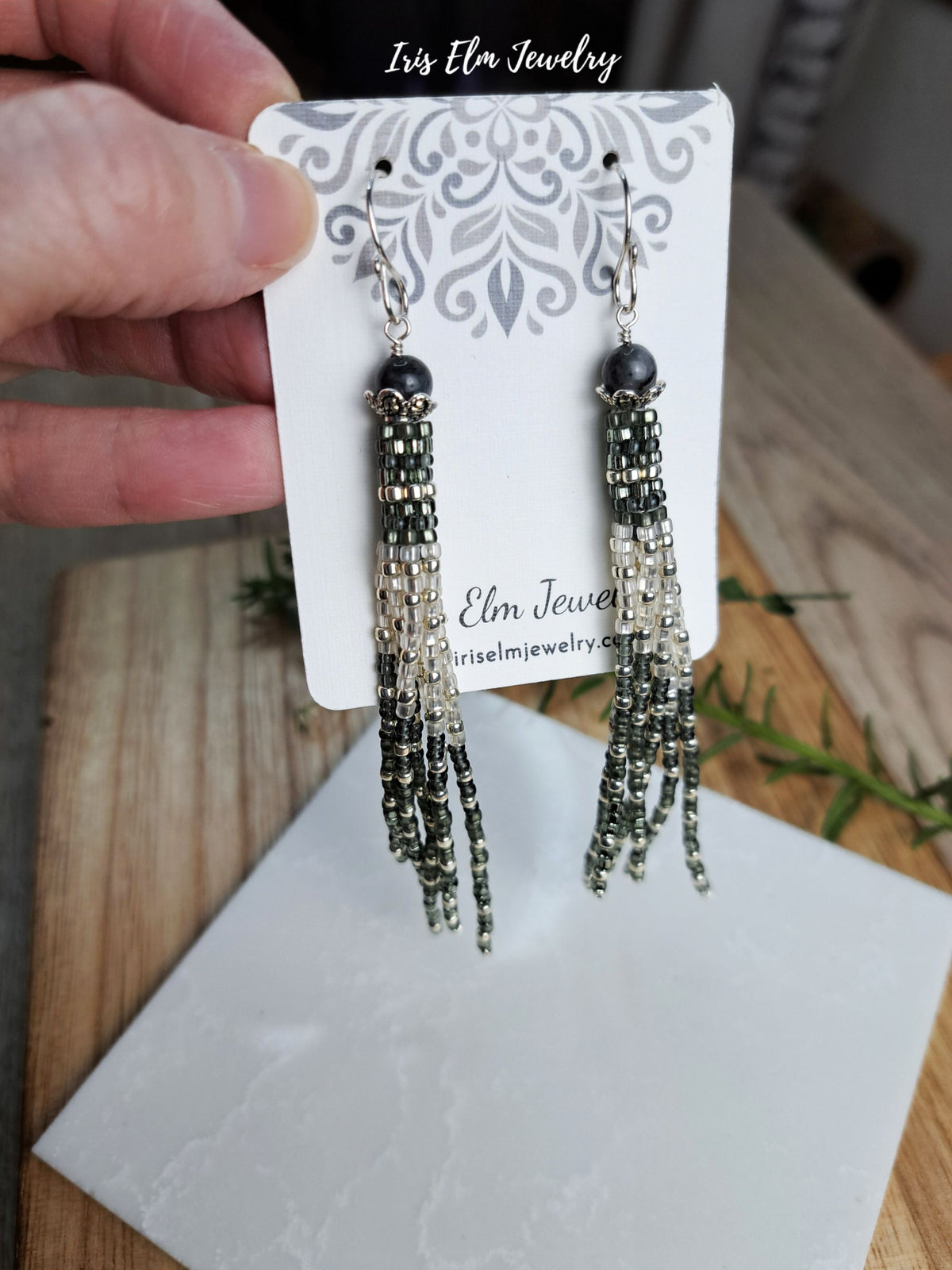A pair of long beaded tassel earrings displayed on a white branded card, held by a hand over a wooden surface. The earrings have dark metallic beads and silver-toned beaded strands.