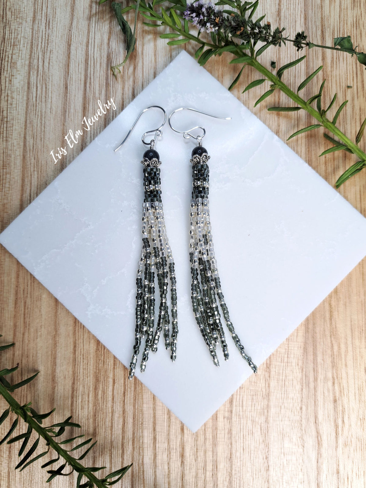 A pair of long beaded tassel drop earrings with silver fishhook wires, displayed on a white square card over a light wood background with green sprigs.