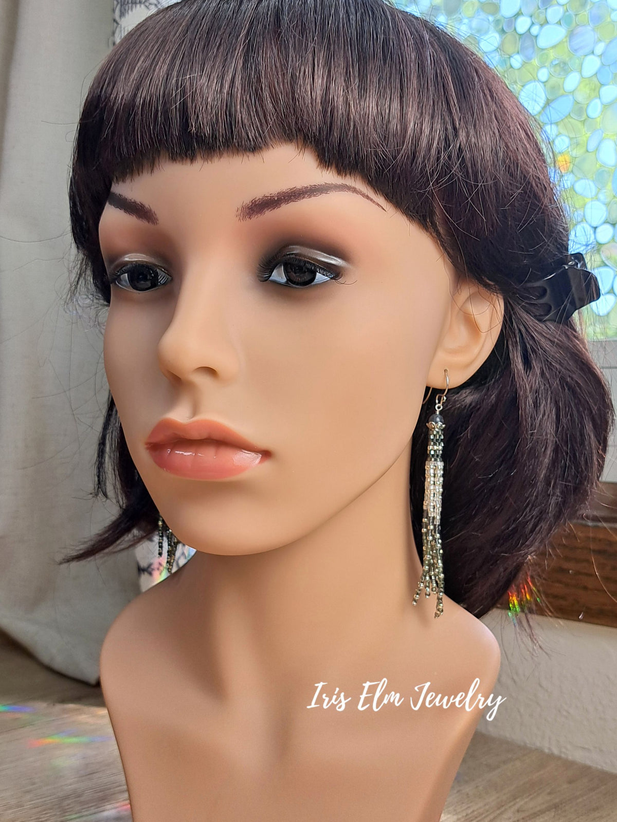 A mannequin bust wearing a short dark brown wig with straight bangs and dangling beaded earrings. The mannequin is photographed indoors with a softly blurred background; a watermark reading 'Iris Elm Jewelry' appears near the lower right.