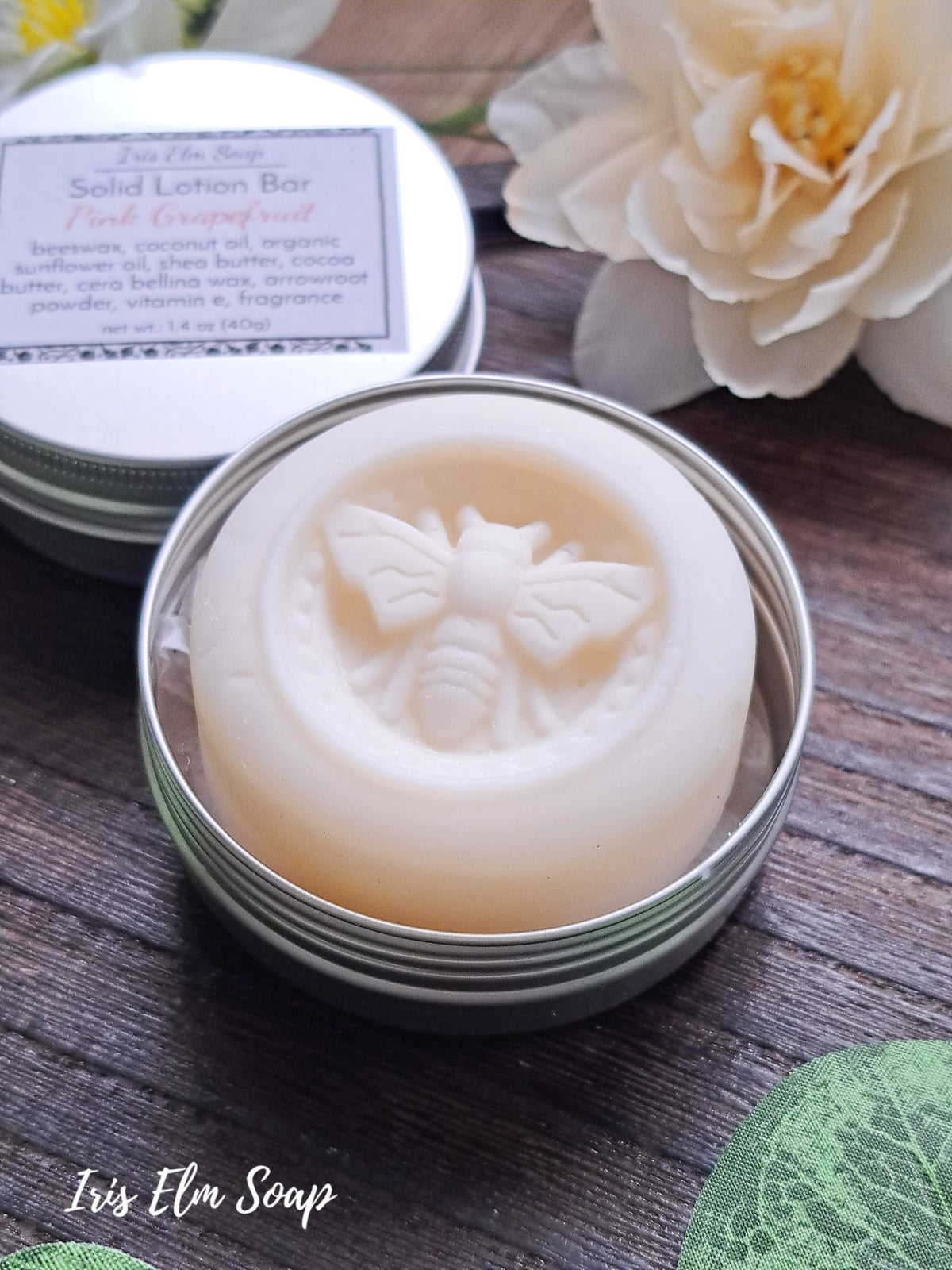 Solid lotion bar with bee design in a tin container on a wooden surface with flowers in the background.