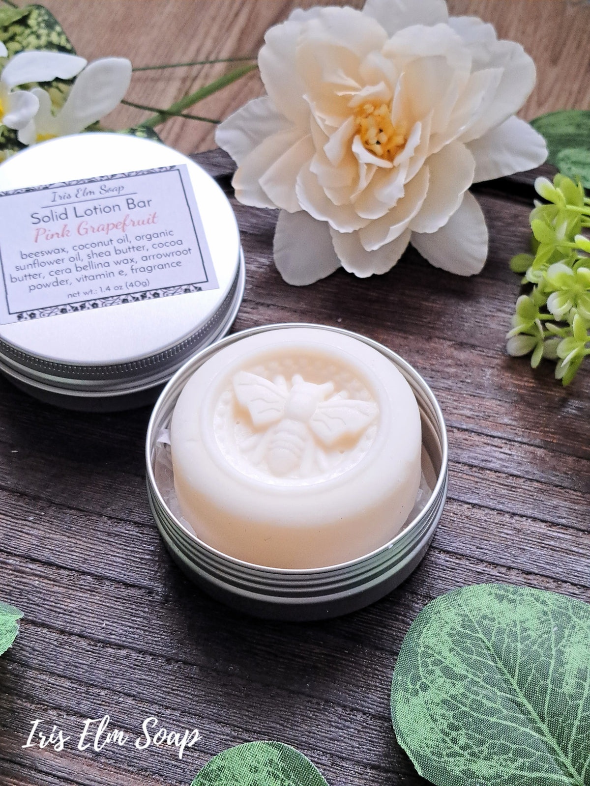 Solid lotion bar in a tin with a bee design, surrounded by flowers and leaves on a wooden surface.