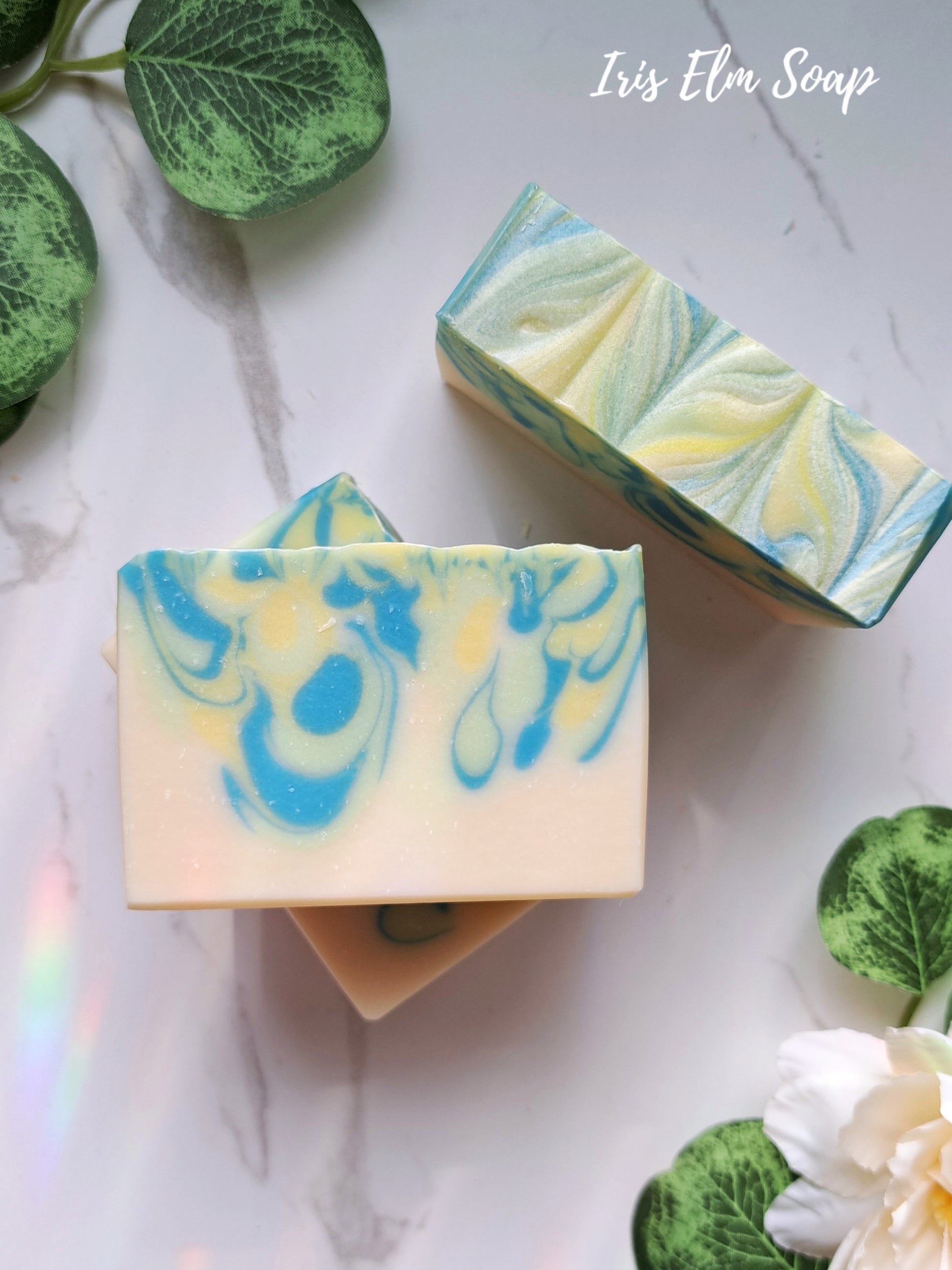 Two bars of Iris Elm Soap with a marbled design on a light background with green leaves and a white flower.