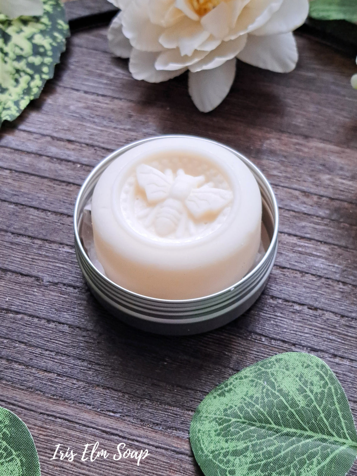 beeswax solid lotion bar with bee design in a metal tin on a wooden surface with floral decorations.