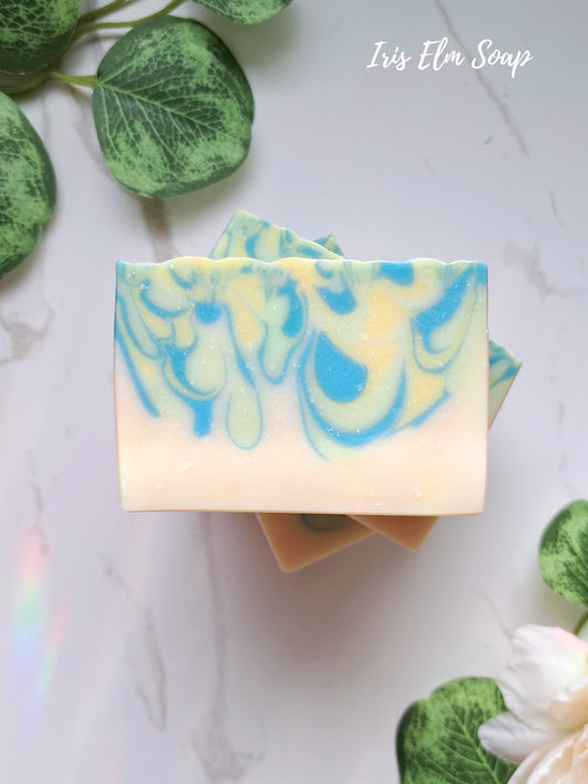 Two swirled white tea and ginger handmade soap bars with blue and green patterns on a white background with leaves.