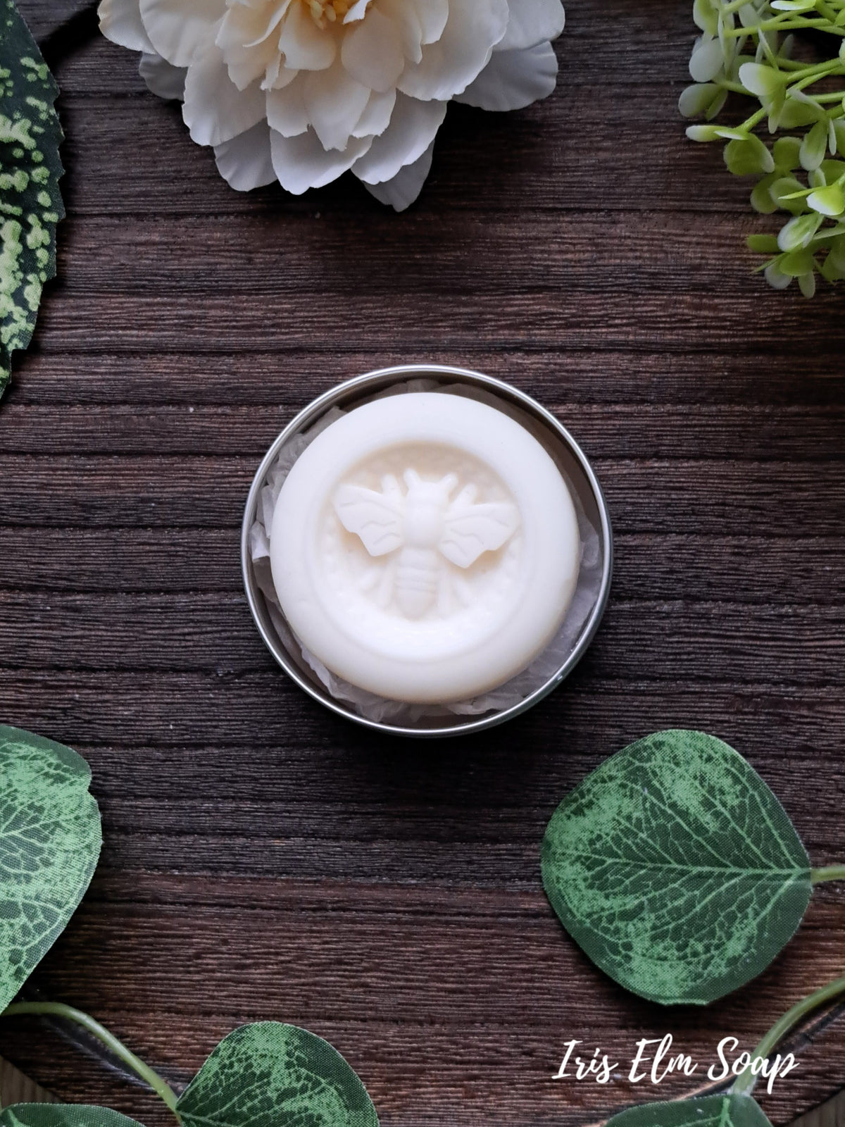 White lotion bar with bee design in a silver container on a wooden surface with green leaves and flowers.