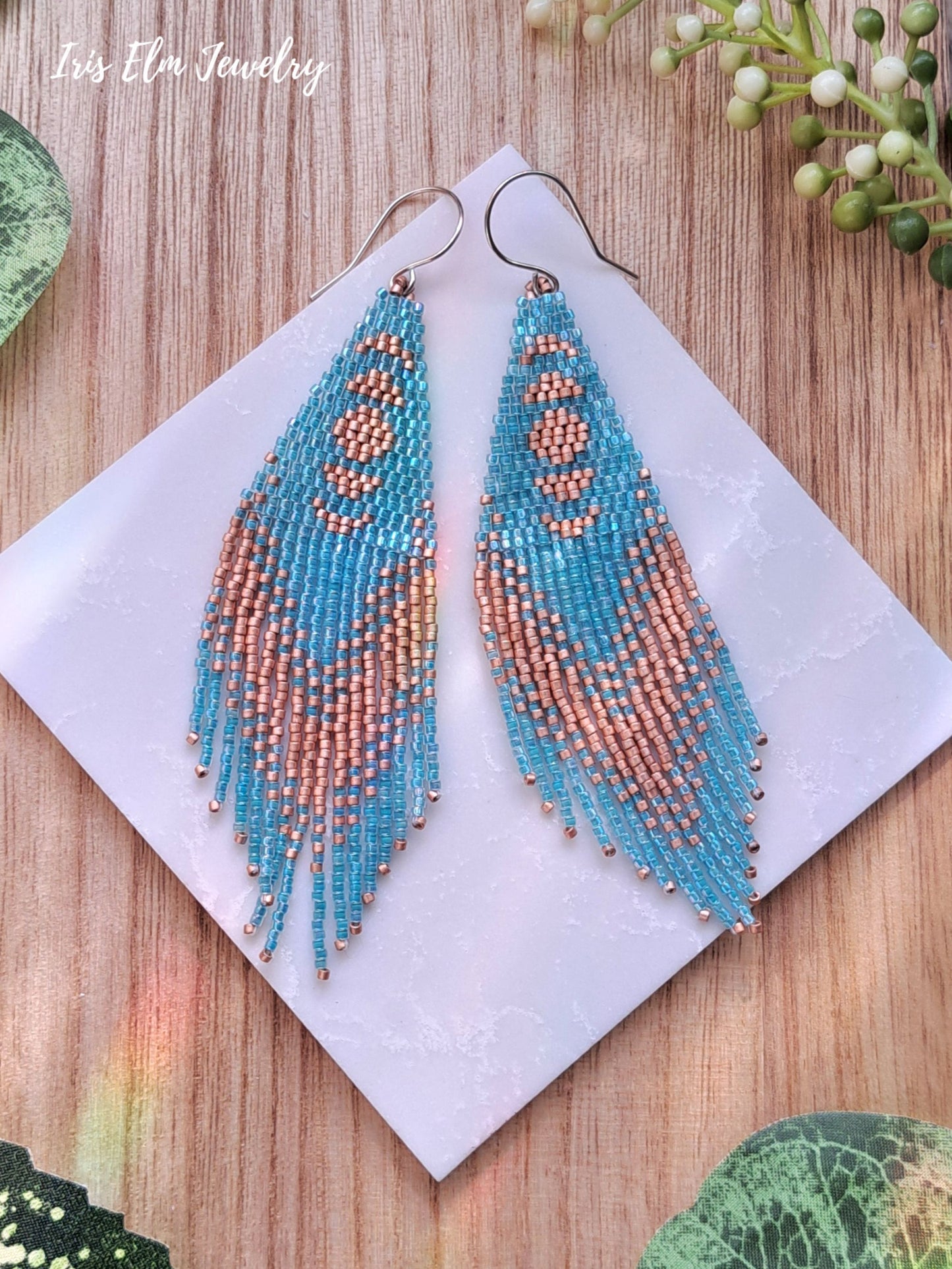Beadwork aqua and copper seed bead moon phase fringe earrings on a white card with 'Iris Elm Jewelry' branding, on a wooden surface.