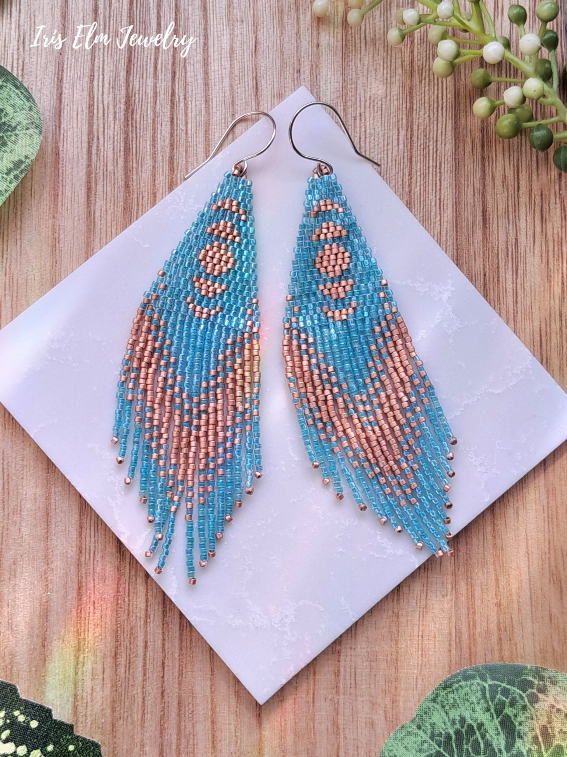 Beadwork aqua and copper seed bead moon phase fringe earrings on a white card with 'Iris Elm Jewelry' branding, on a wooden surface.