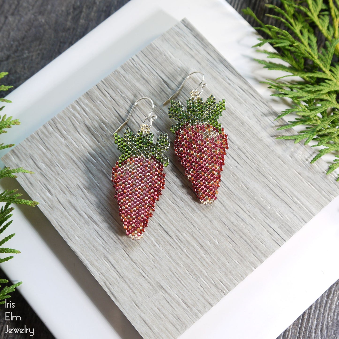 Beaded red strawberry-shaped glass seed bead earrings on a wooden surface with greenery around.
