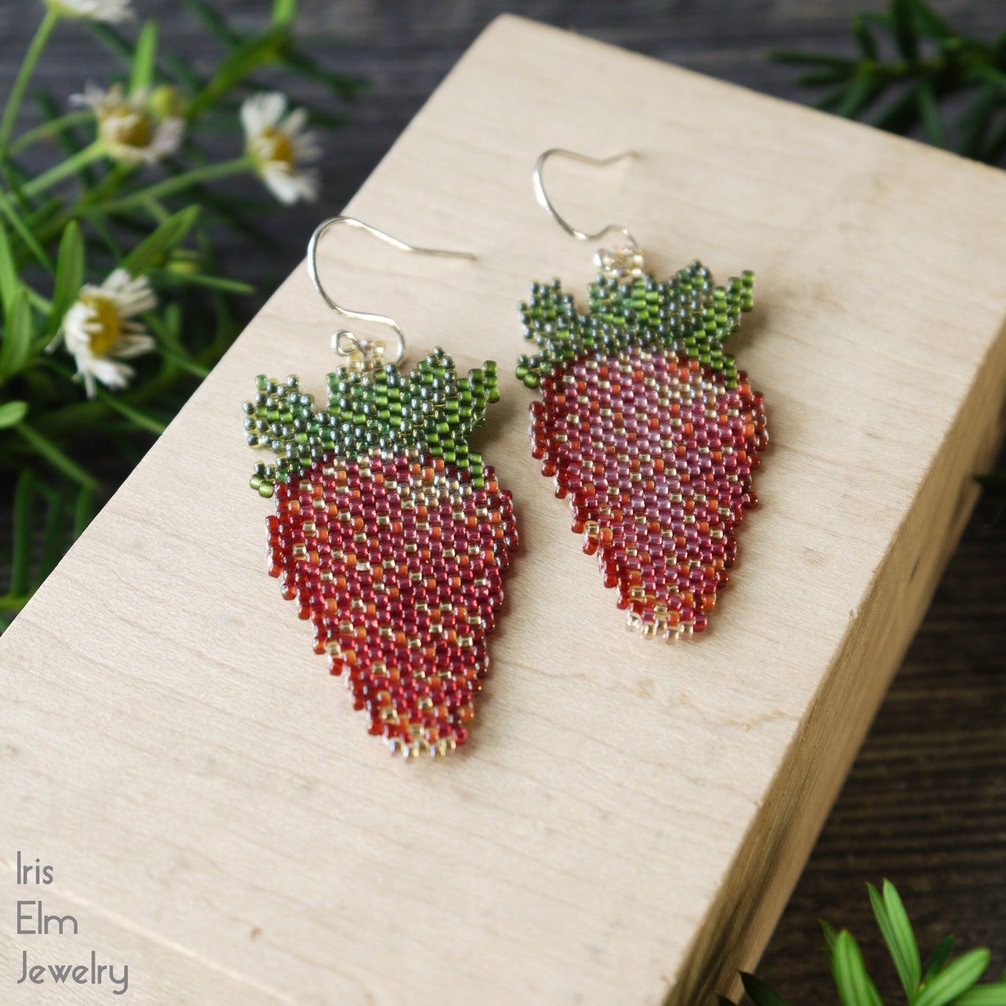 red strawberry-shaped beaded seed bead earrings on a wooden block with plants in the background