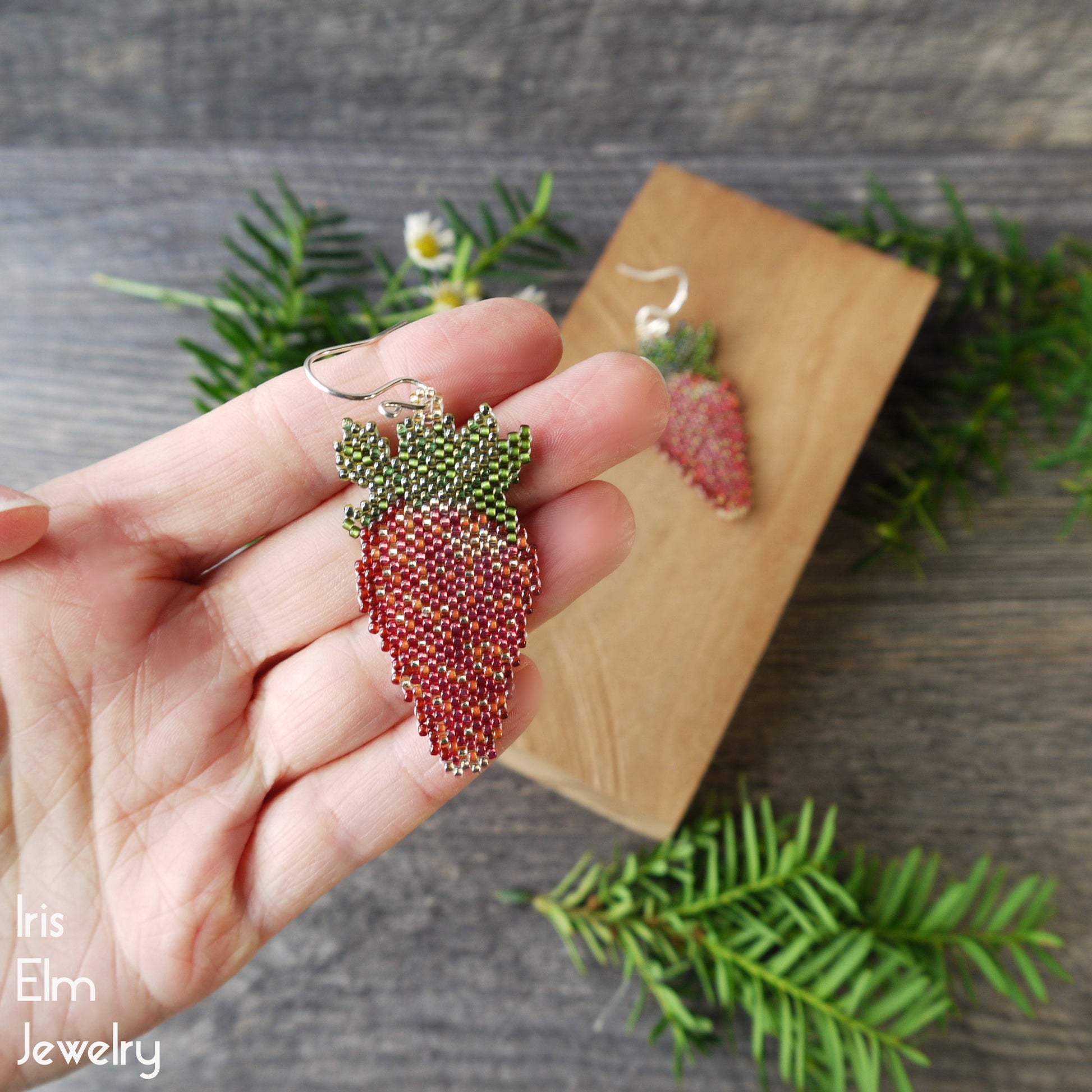 Hand holding a beaded strawberry-shaped earring with a wooden card and green leaves in the background.