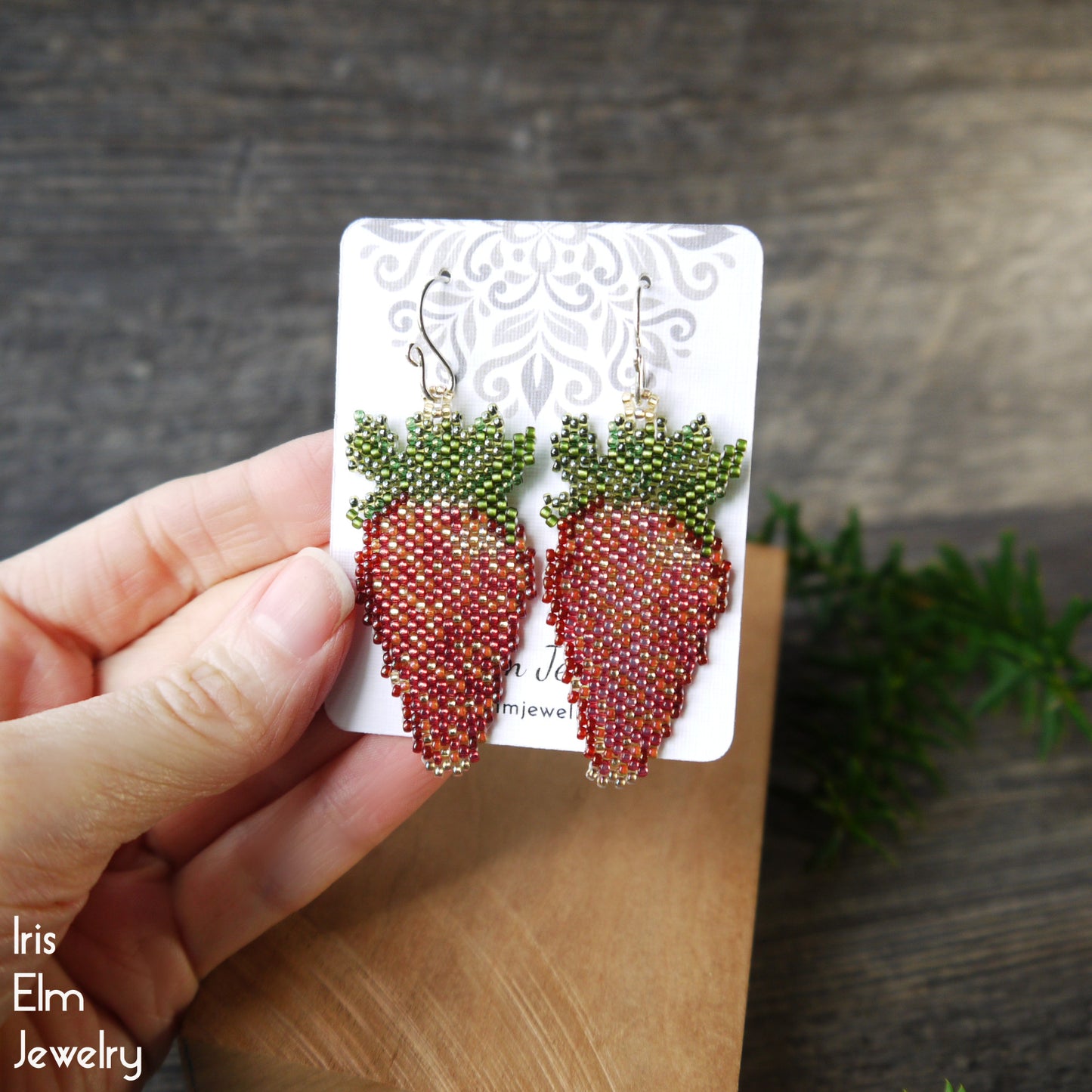 Hand holding a card with beaded strawberry earrings on a wooden surface.