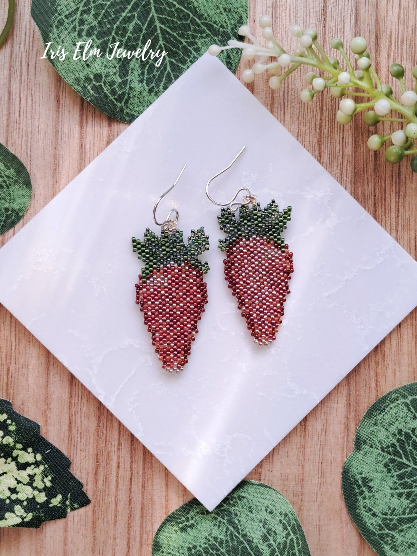 Strawberry-shaped beaded earrings on a white tile with green leaves and flowers in the background.