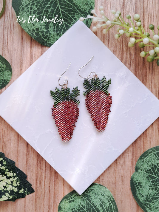 Strawberry-shaped beaded earrings on a white tile with green leaves and flowers in the background.