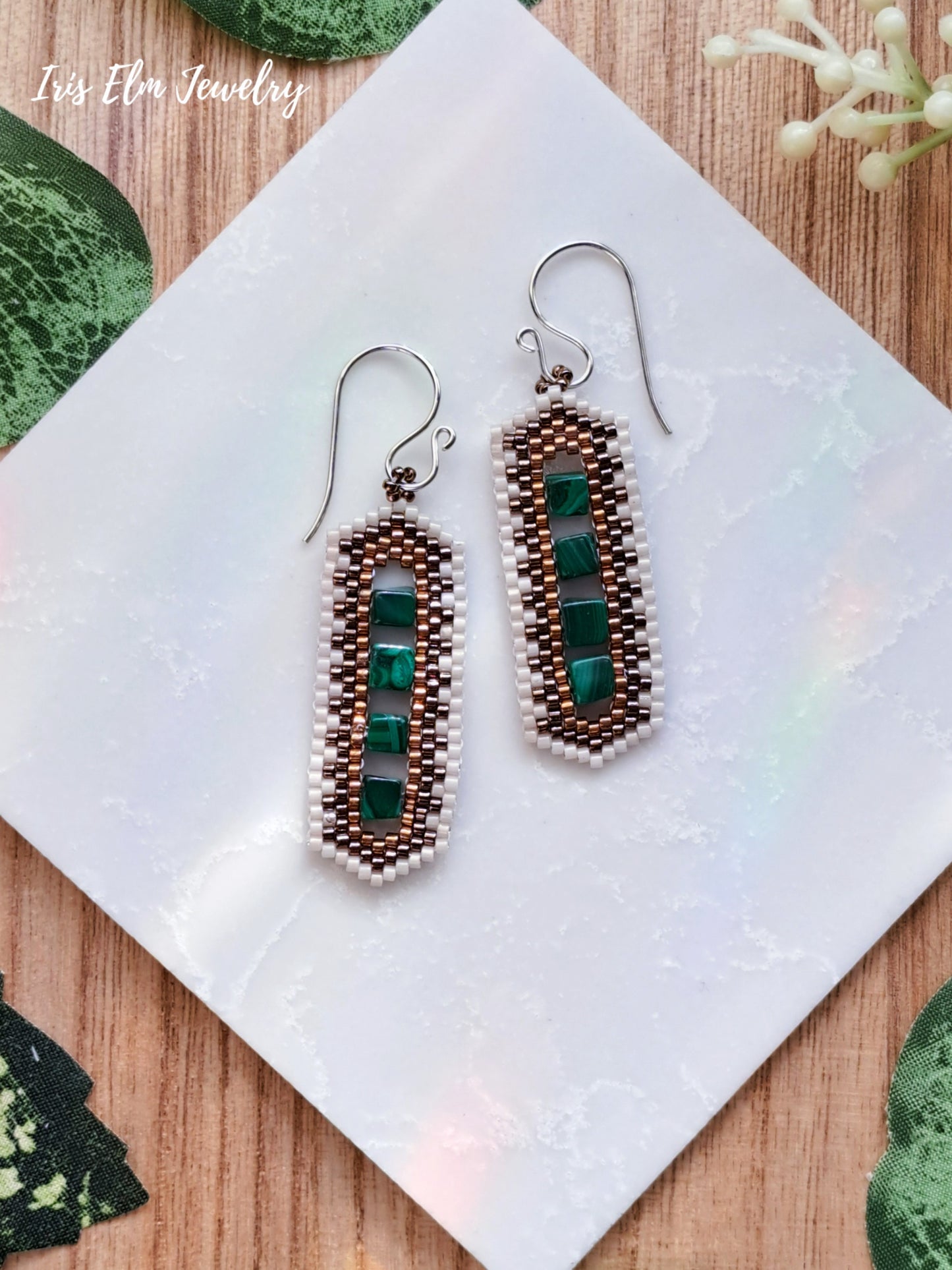 Pair of beaded earrings with green malachite stones on a white cloth with wooden surface and green leaves in the background.
