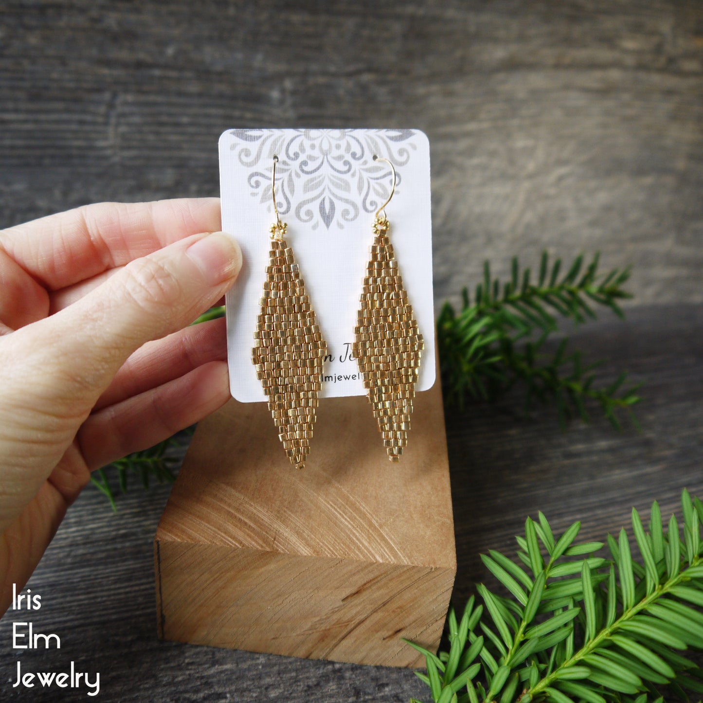 Gold beadwork glass bead earrings on a wooden stand with a hand holding the stand, against a natural background.