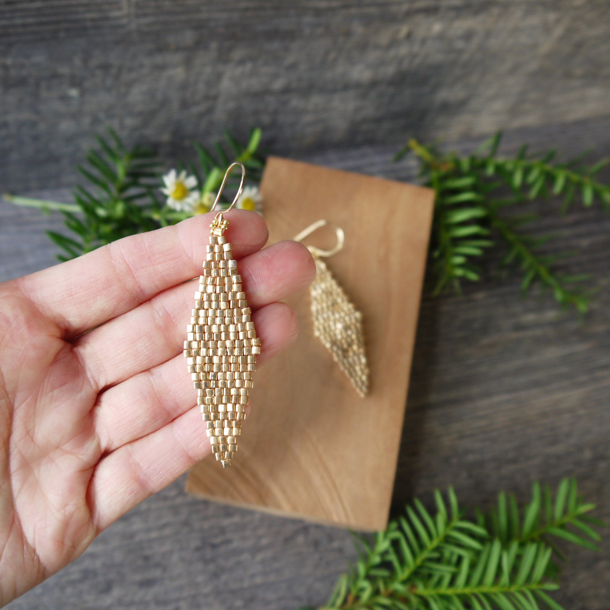 Gold beaded glass diamond woven earrings held in a hand with a wooden block and greenery in the background.