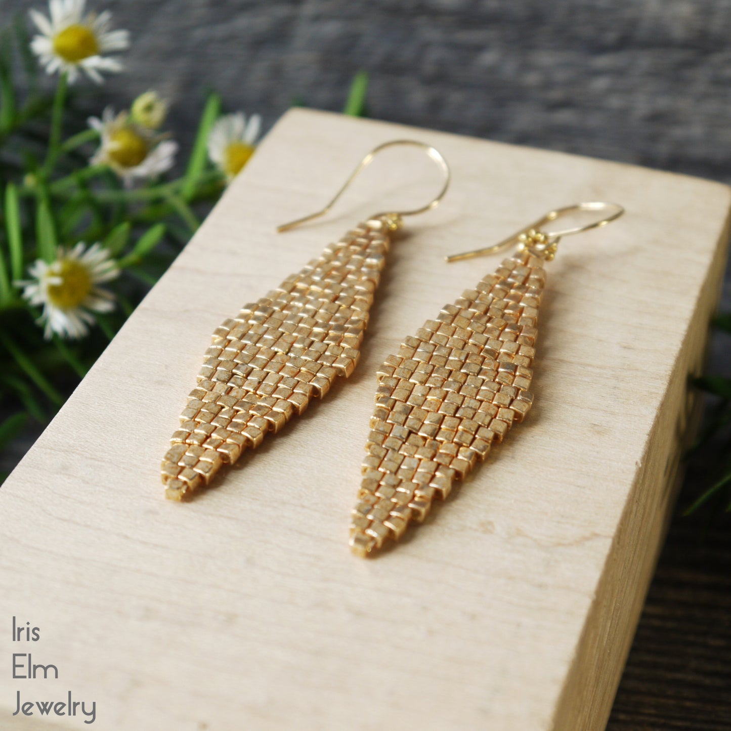 Gold beaded diamond shape woven handmade earrings on a wooden block with flowers in the background, featuring 'Iris Elm Jewelry' branding.
