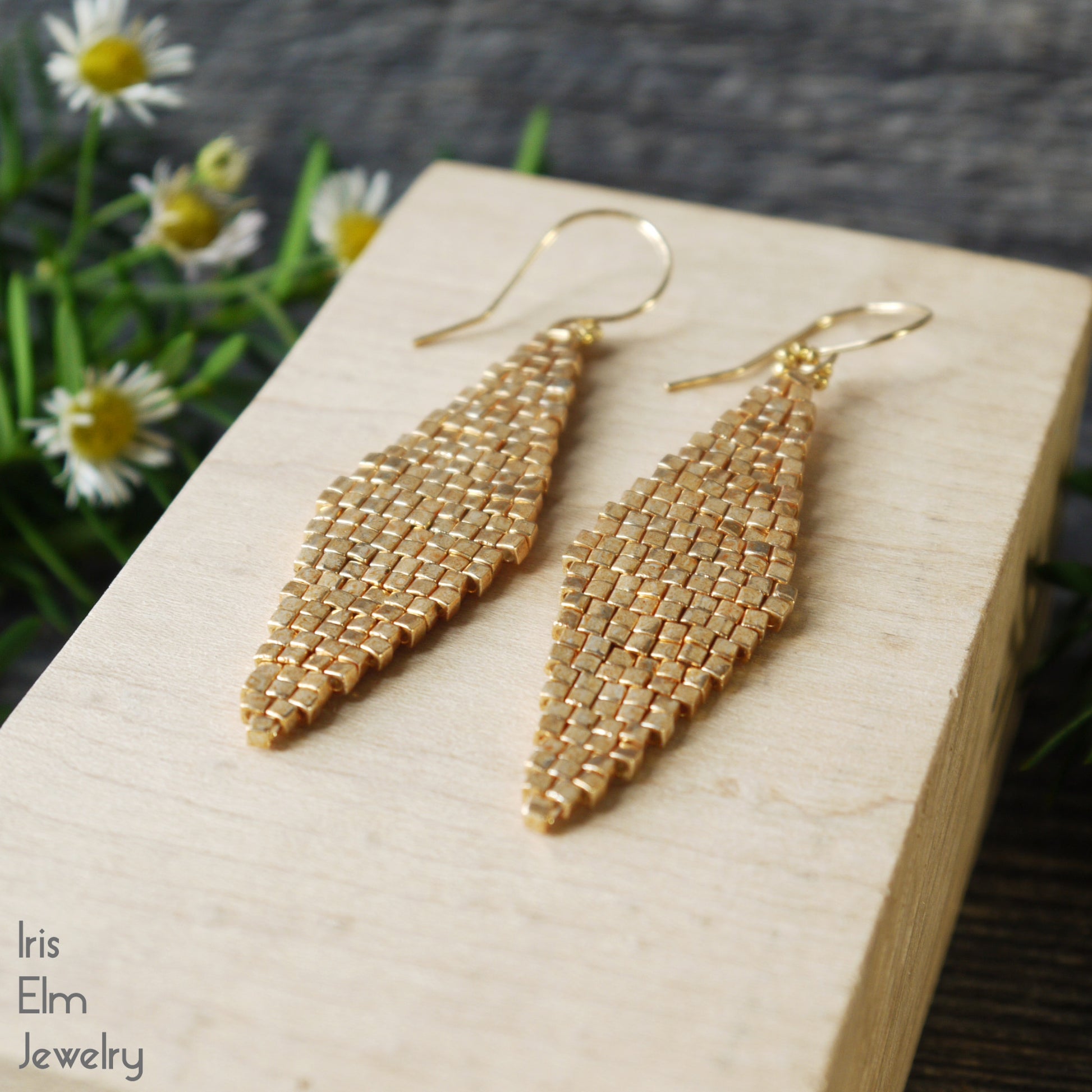 Gold beaded diamond shape woven handmade earrings on a wooden block with flowers in the background, featuring 'Iris Elm Jewelry' branding.