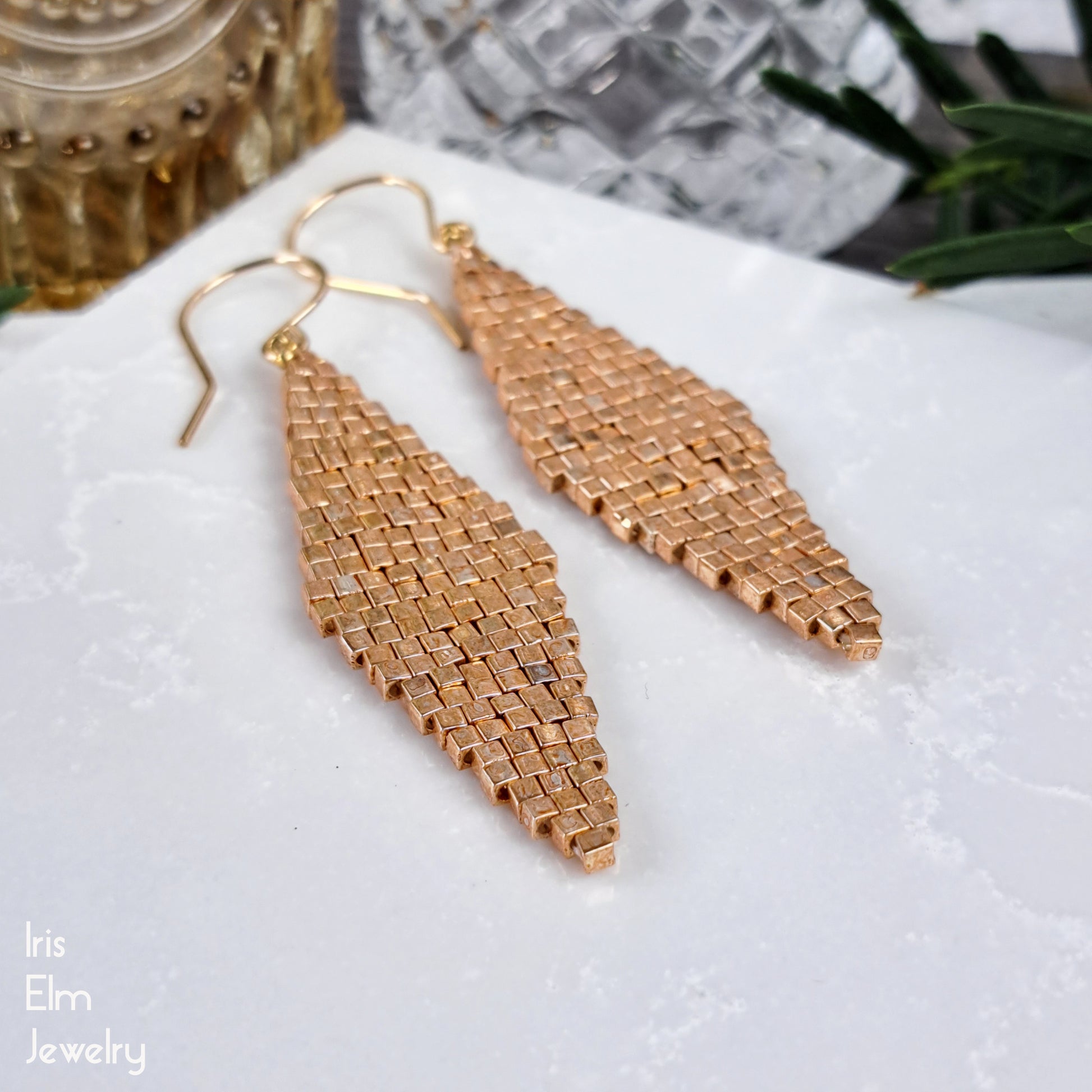 gold colored glass woven earrings on a white surface with 'Iris Elm Jewelry' branding.