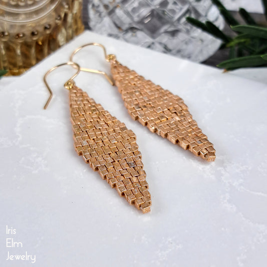 gold colored glass woven earrings on a white surface with 'Iris Elm Jewelry' branding.