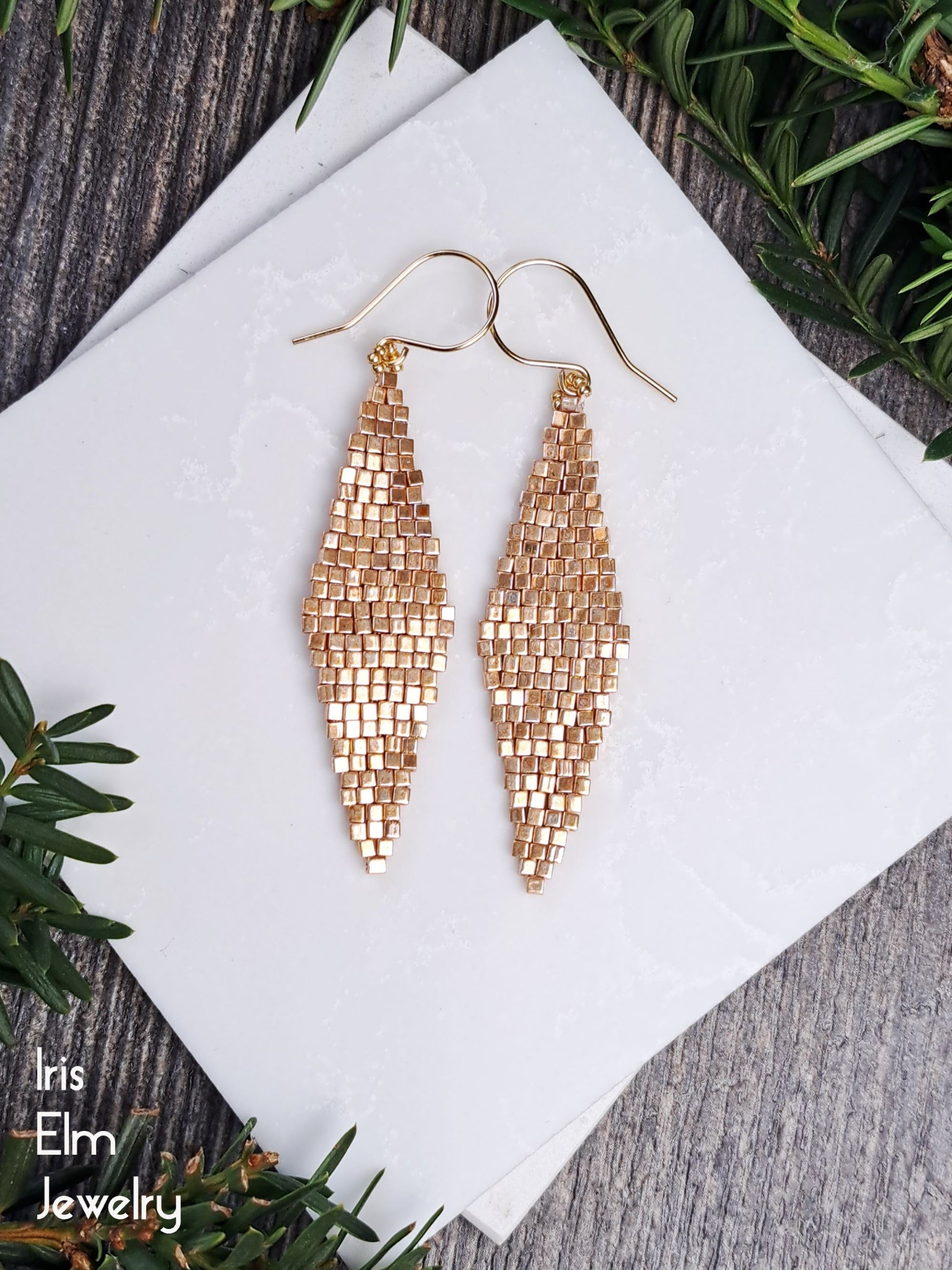 Pair of gold seed bead long diamond shape earrings on a white napkin with greenery around.