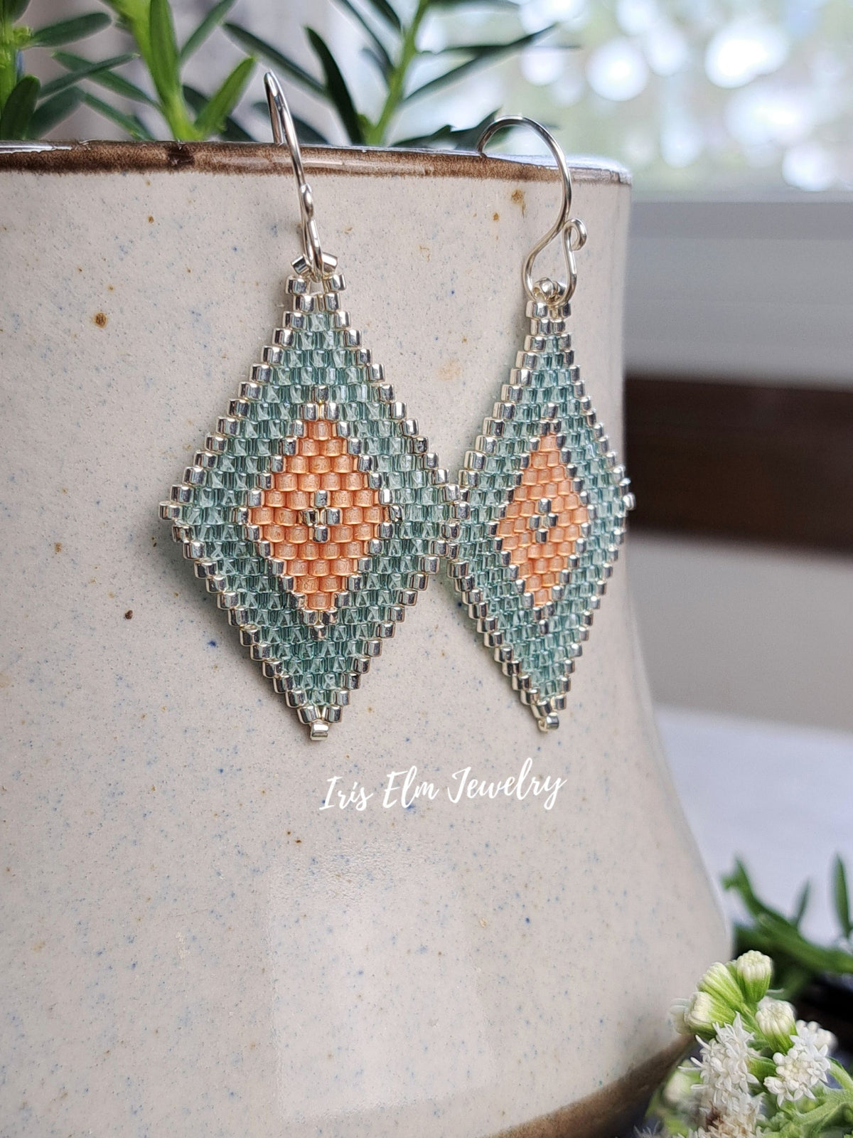 A pair of handmade beaded dangle earrings with a geometric diamond design in aqua green, peach, and silver, hanging from a ceramic pot with a watermark reading 'Iris Elm Jewelry'.