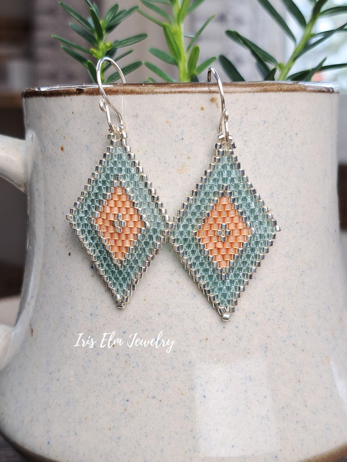 A pair of handmade beaded dangle earrings with a geometric diamond pattern in aqua green and peach, hanging from silver hooks in front of a speckled ceramic mug with green leaves in the background.