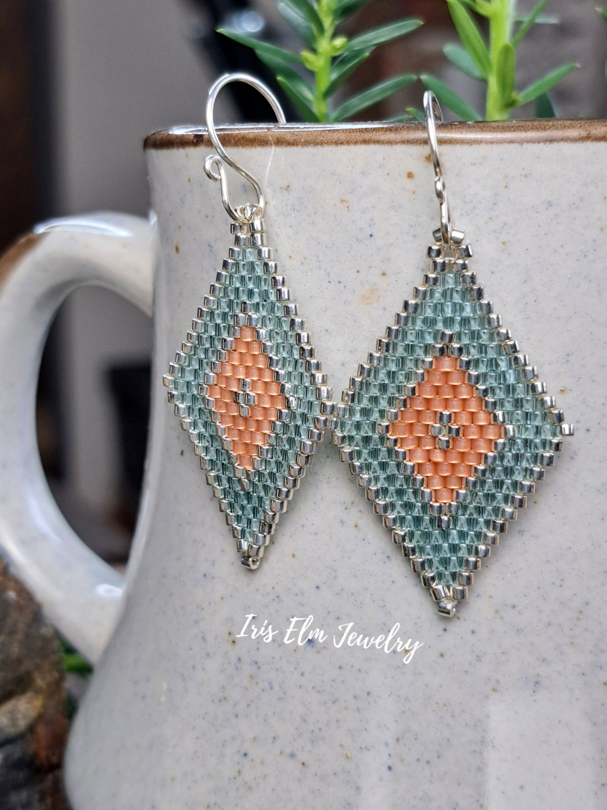 A pair of handmade beaded dangle earrings with a silver-tone metal frame and hooks, featuring a geometric pattern in aqua green and peach. The earrings are displayed hanging in front of a speckled ceramic mug with green plants in the background.
