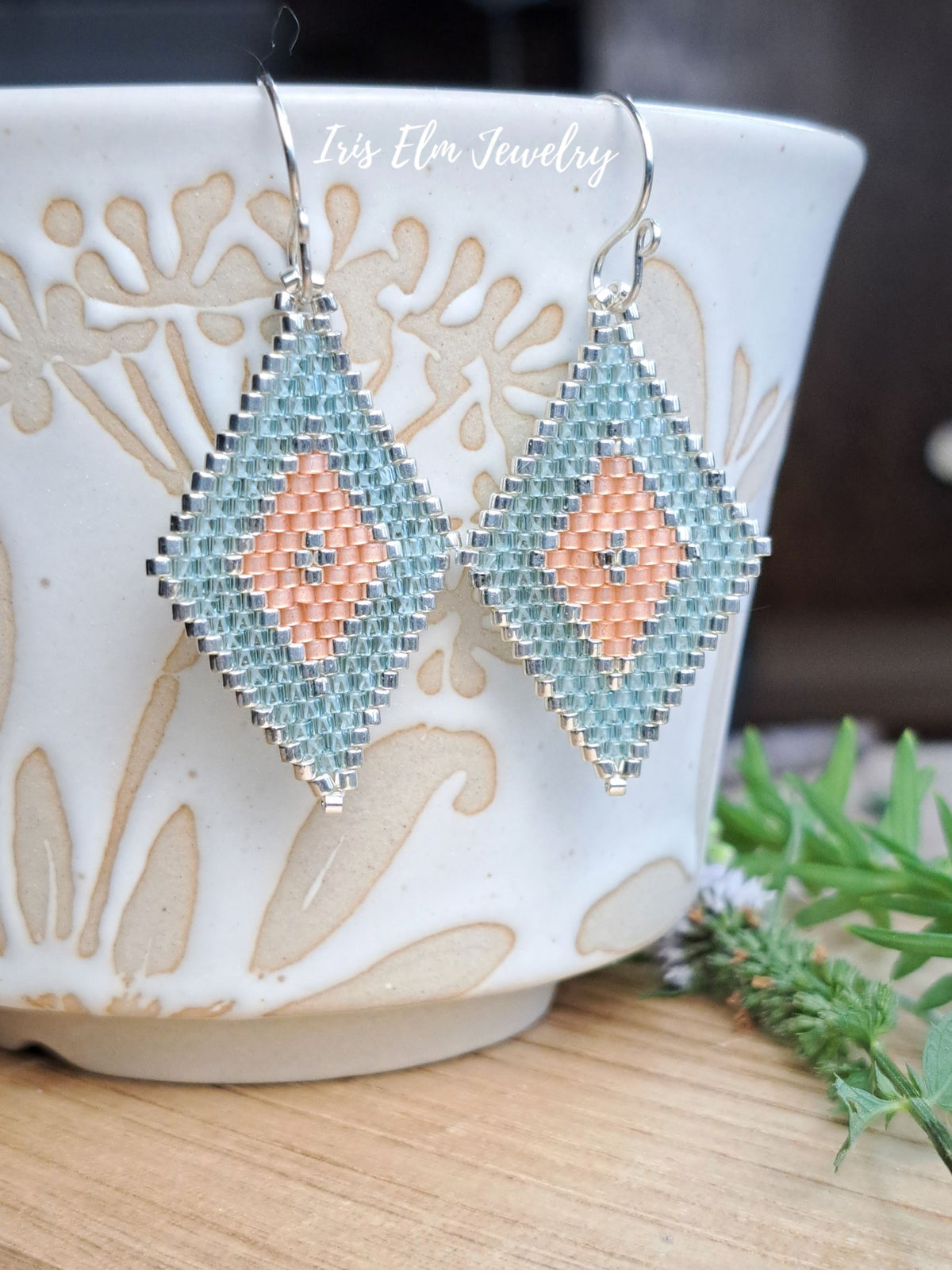 A pair of handmade beaded dangle earrings with a silver-toned metal frame, featuring a geometric pattern in light blue and peach. The earrings are displayed on a white ceramic cup with beige floral patterns, with a small sprig of green herbs on a wooden surface in the foreground.