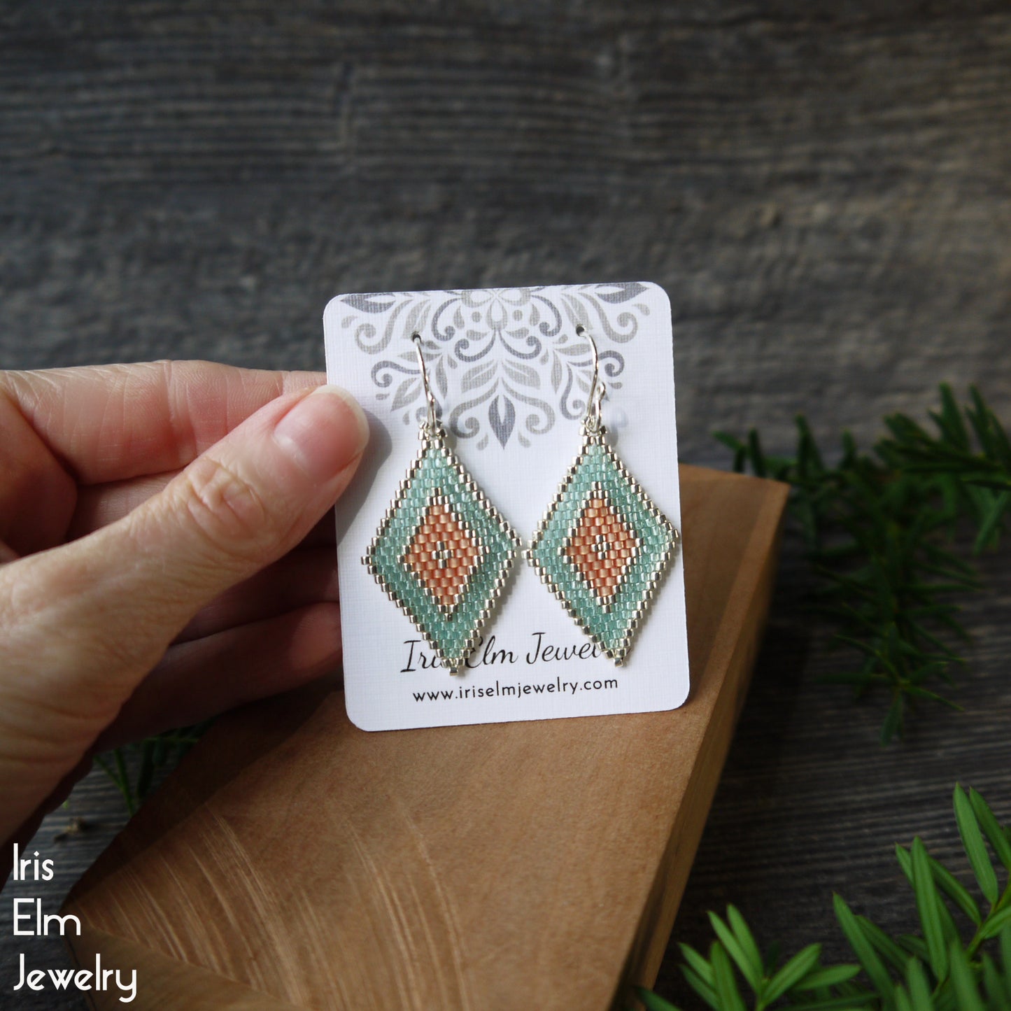 Hand holding a pair of diamond shape seed bead earrings on an Iris Elm Jewelry card against a wooden surface with greenery.