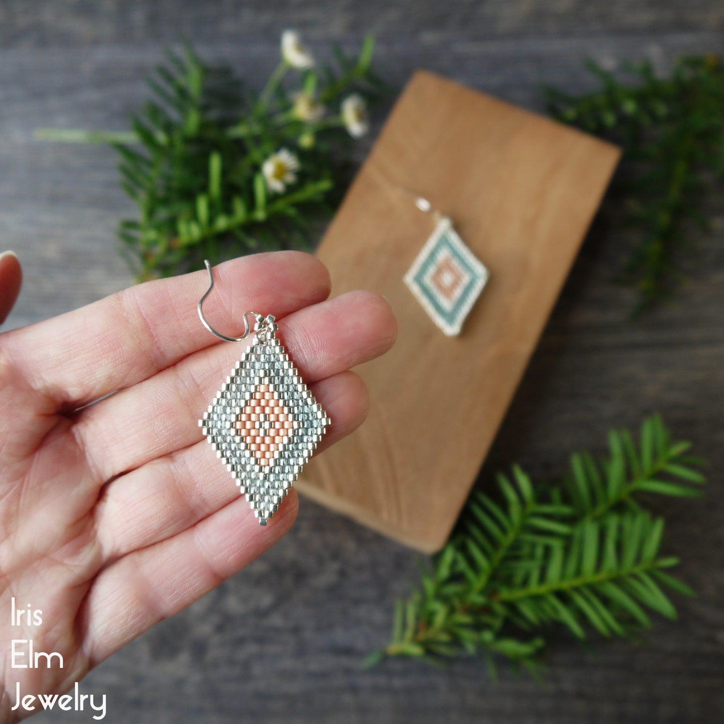 Hand holding a peach and aqua diamond-shaped beaded earring with greenery in the background, featuring 'Iris Elm Jewelry'.