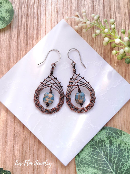 Pair of copper wire woven teardrop earrings with blue aqua terra jasper beads on a white tile with green leaves in the background