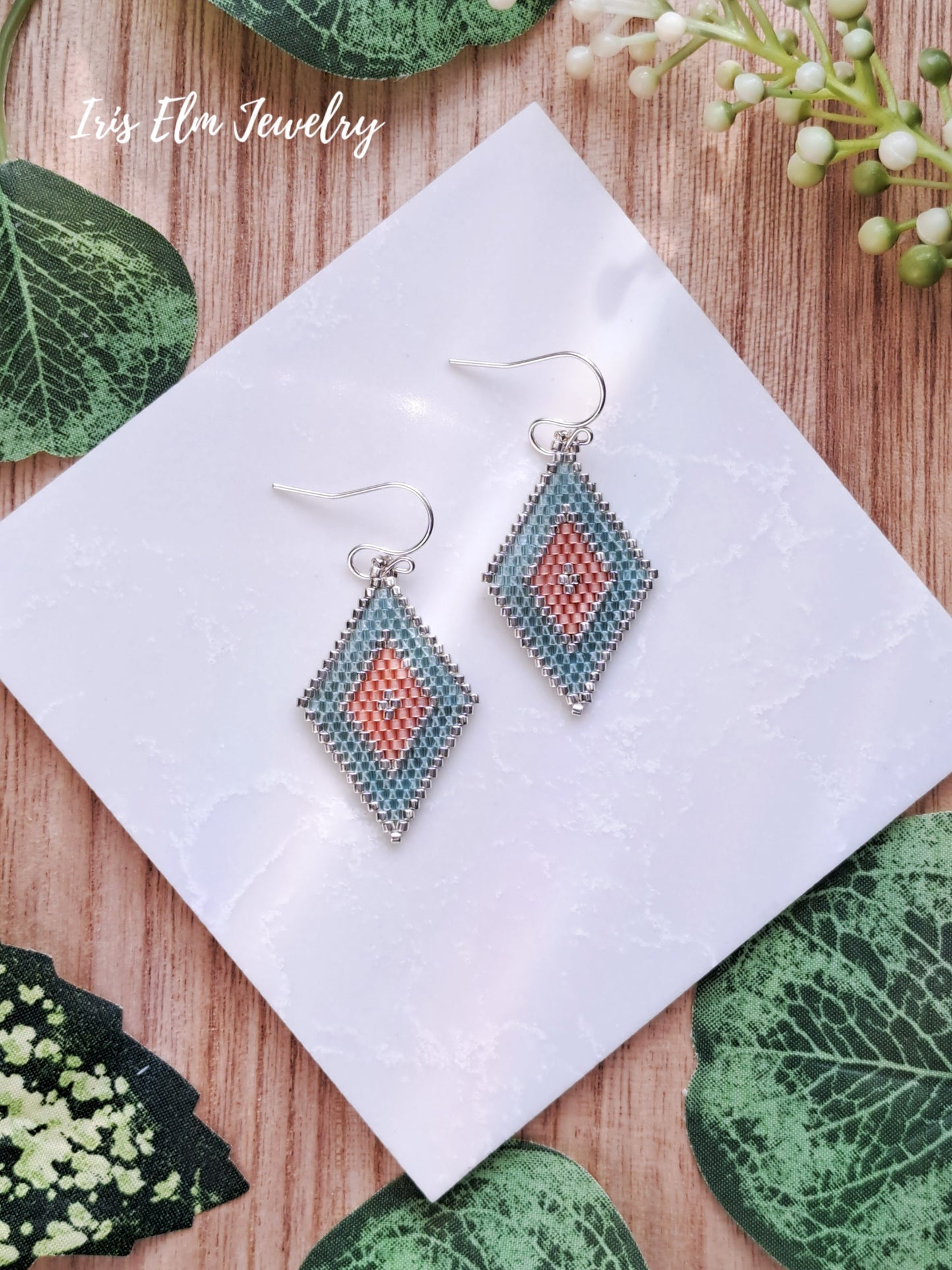 Pair of diamond-shaped peach and aqua southwestern seed bead earrings on a white tile with green leaves in the background, featuring 'Iris Elm Jewelry'.
